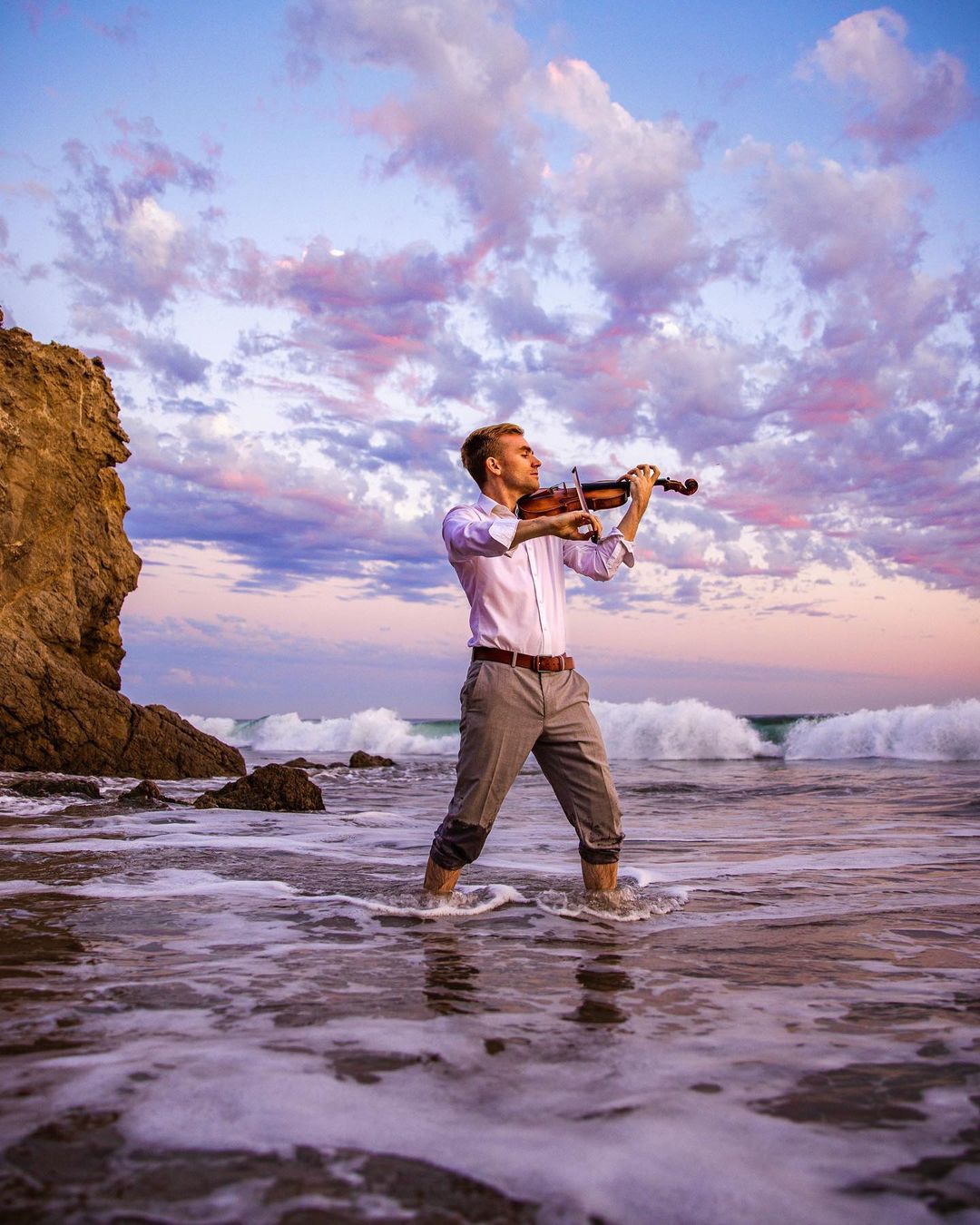 Man playing violin in shallow ocean water during sunset, with clouds and rocky cliff in background.