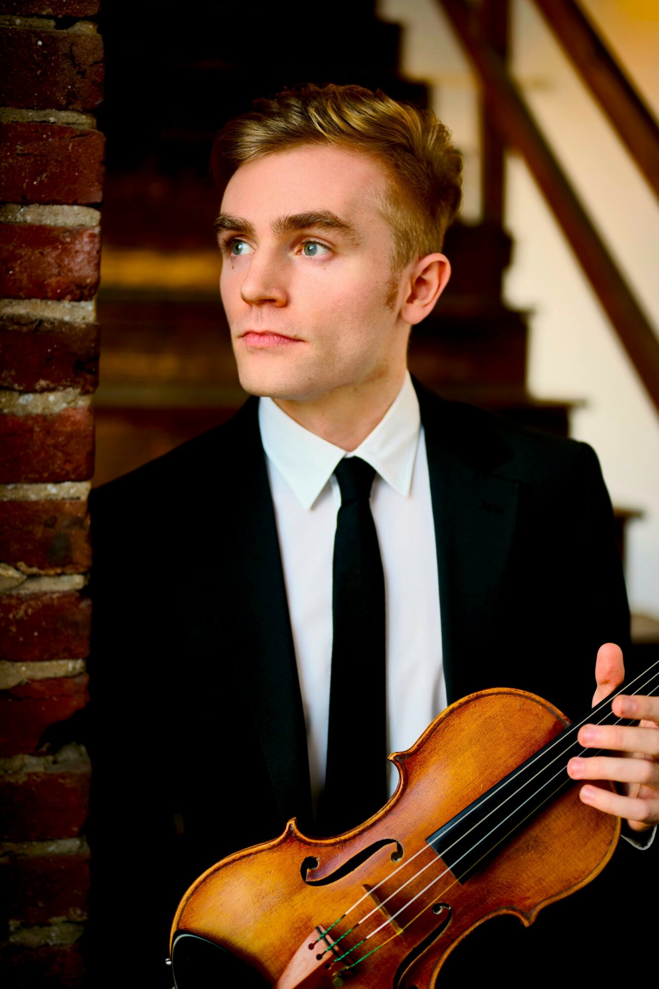 Young man in formal suit holding a violin, standing near a brick wall with stairs in background.