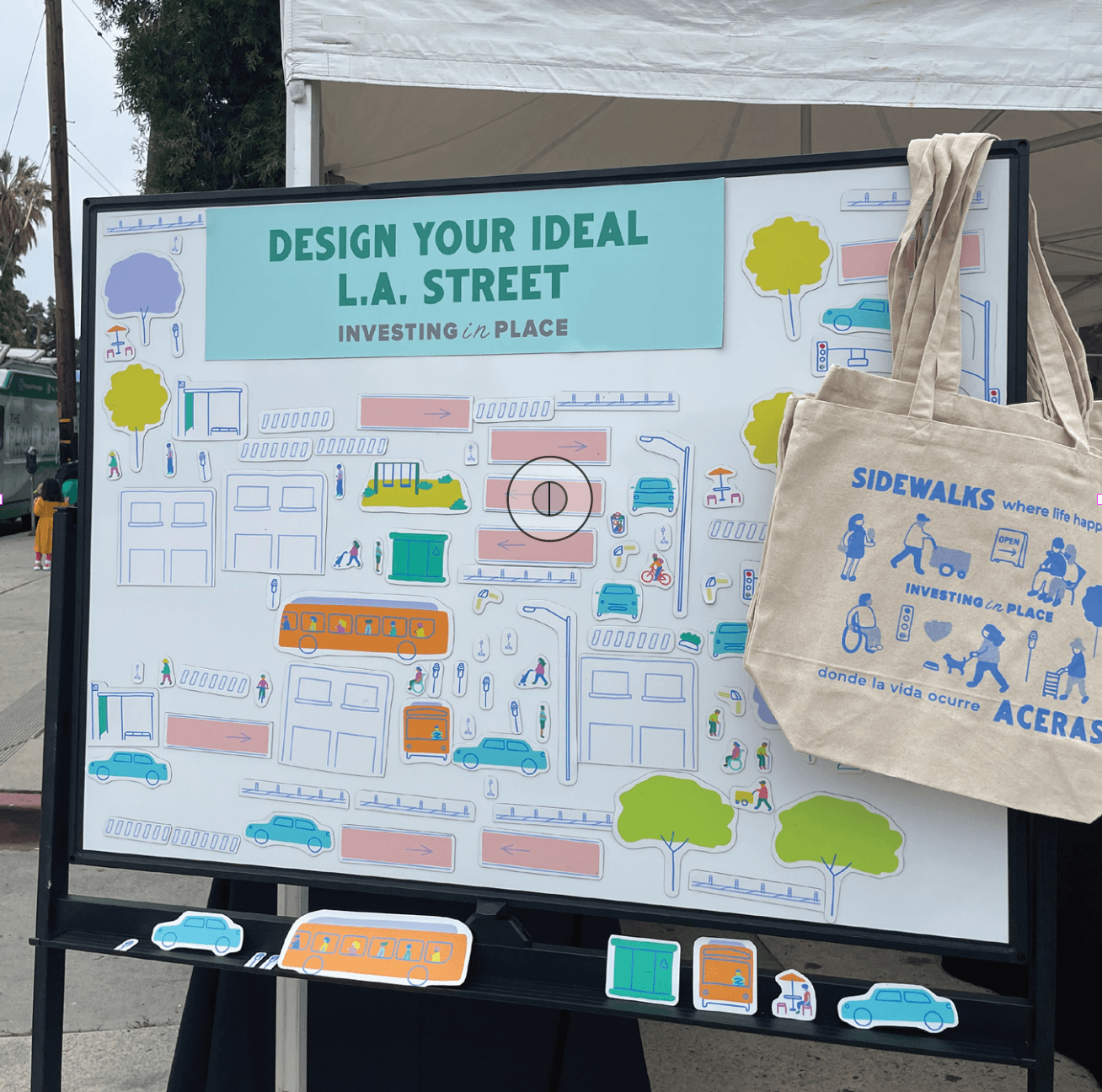 Outdoor display board with a tote bag hanging on the right side, featuring illustrations of city streets and trees, promoting urban design.