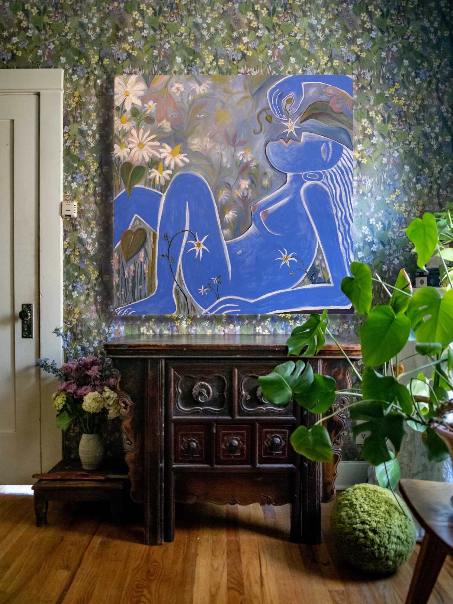 Interior with a colorful abstract painting of a blue figure, floral wallpaper, a dark wooden cabinet, potted plants, and a green moss ball.