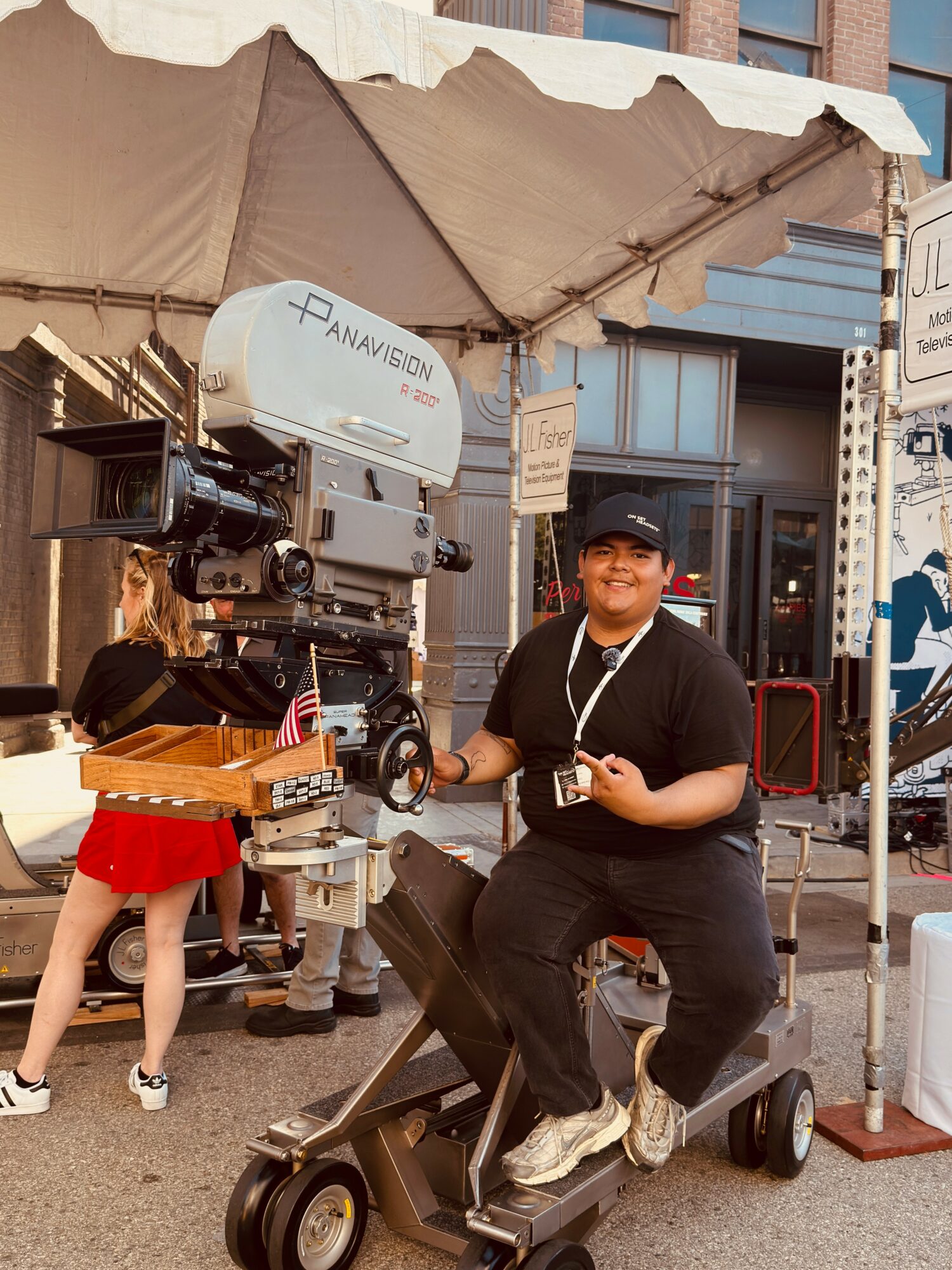 Person sitting on a mobility scooter holding a camera, under a canopy with a film camera setup nearby, on a city street.