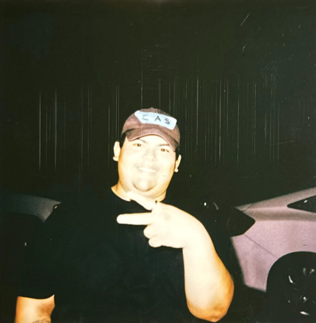 Person wearing a cap and black shirt making a peace sign with hand, standing outdoors at night.