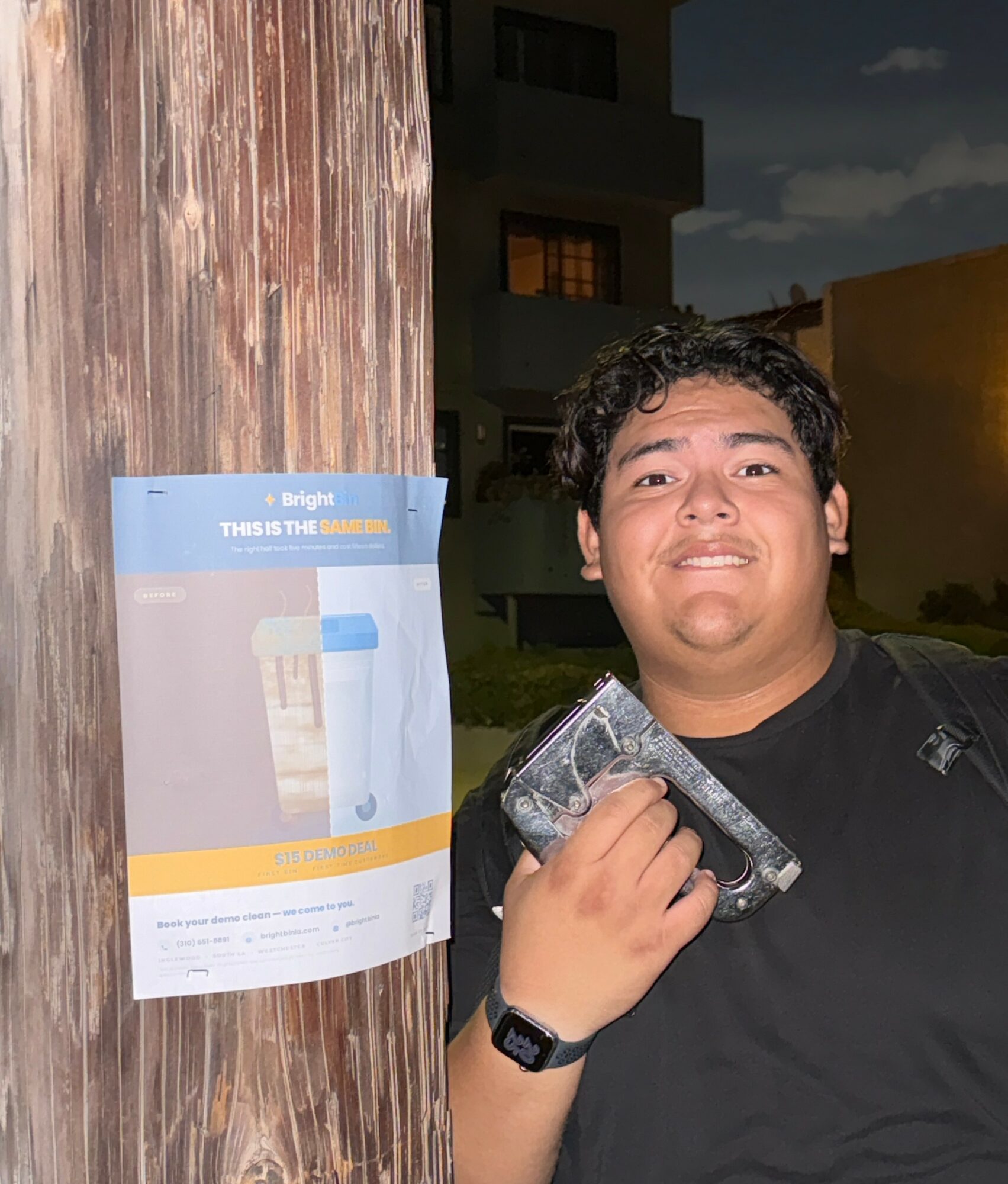 Person holding a camera, standing next to a wooden pole with a poster, outdoors during evening, smiling, wearing a black shirt and watch.