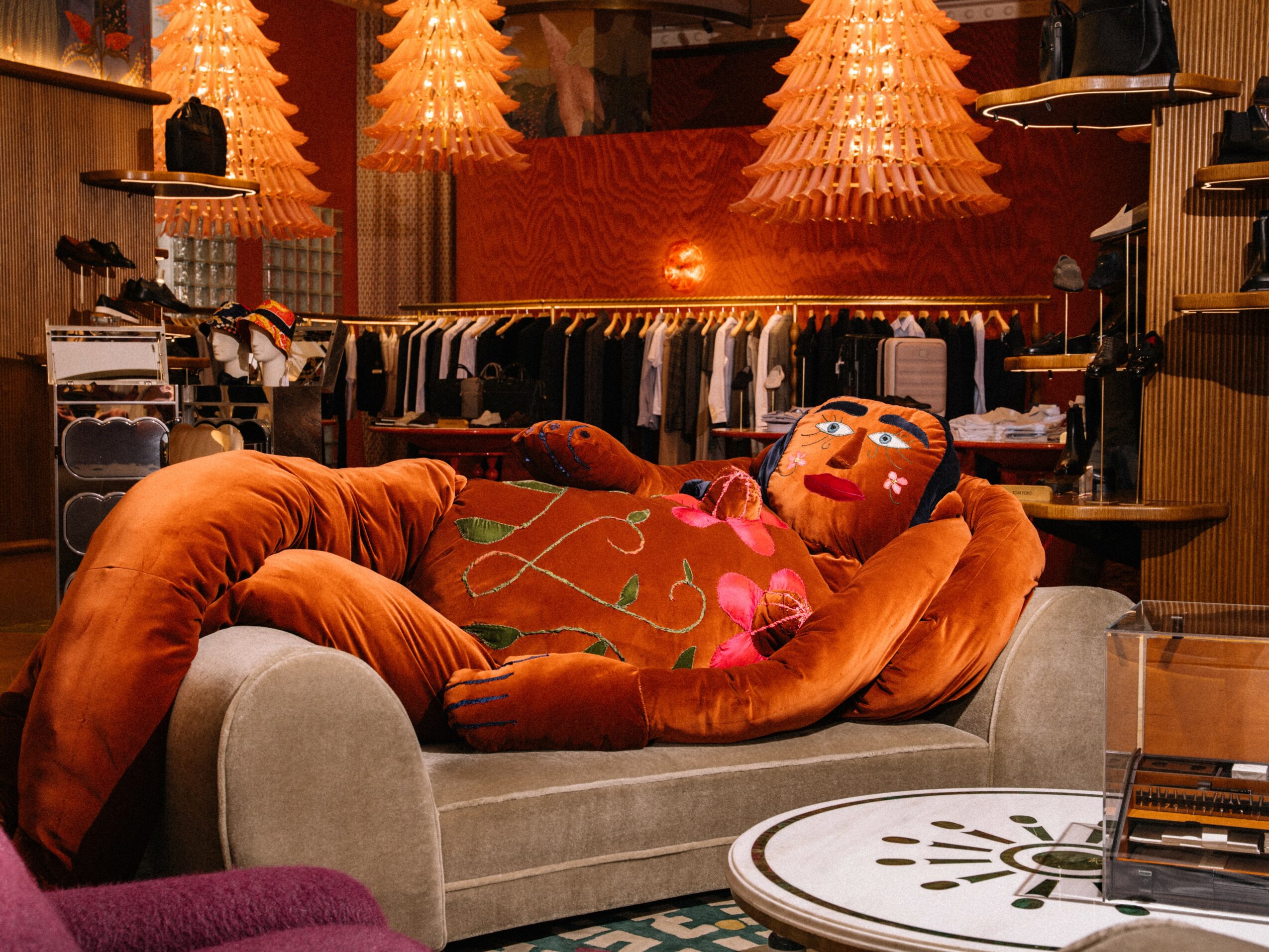 Person lying on a sofa with a colorful pillow, in a clothing store with hanging garments and warm lighting