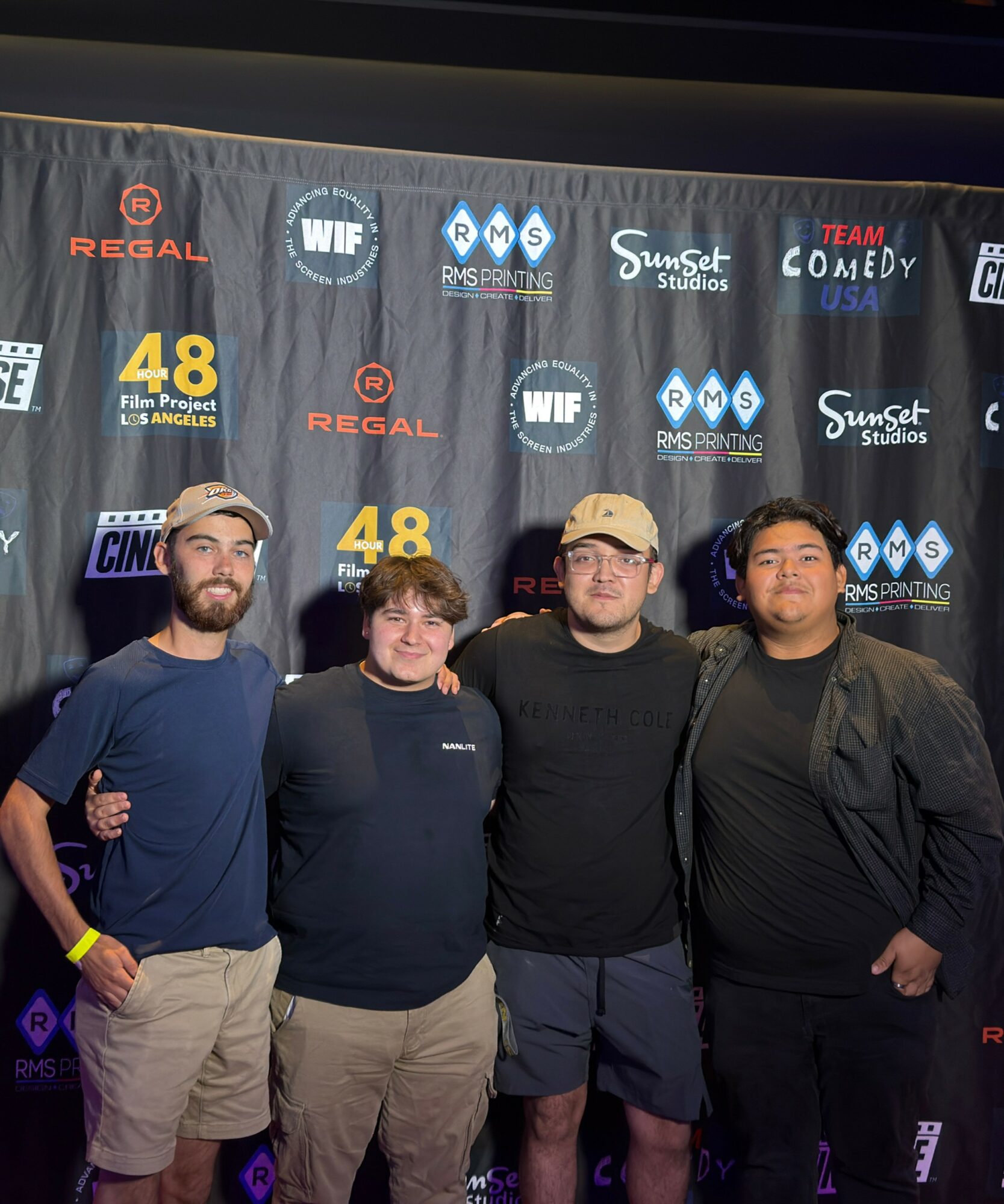 Four people standing in front of a backdrop with logos and text, smiling at the camera.