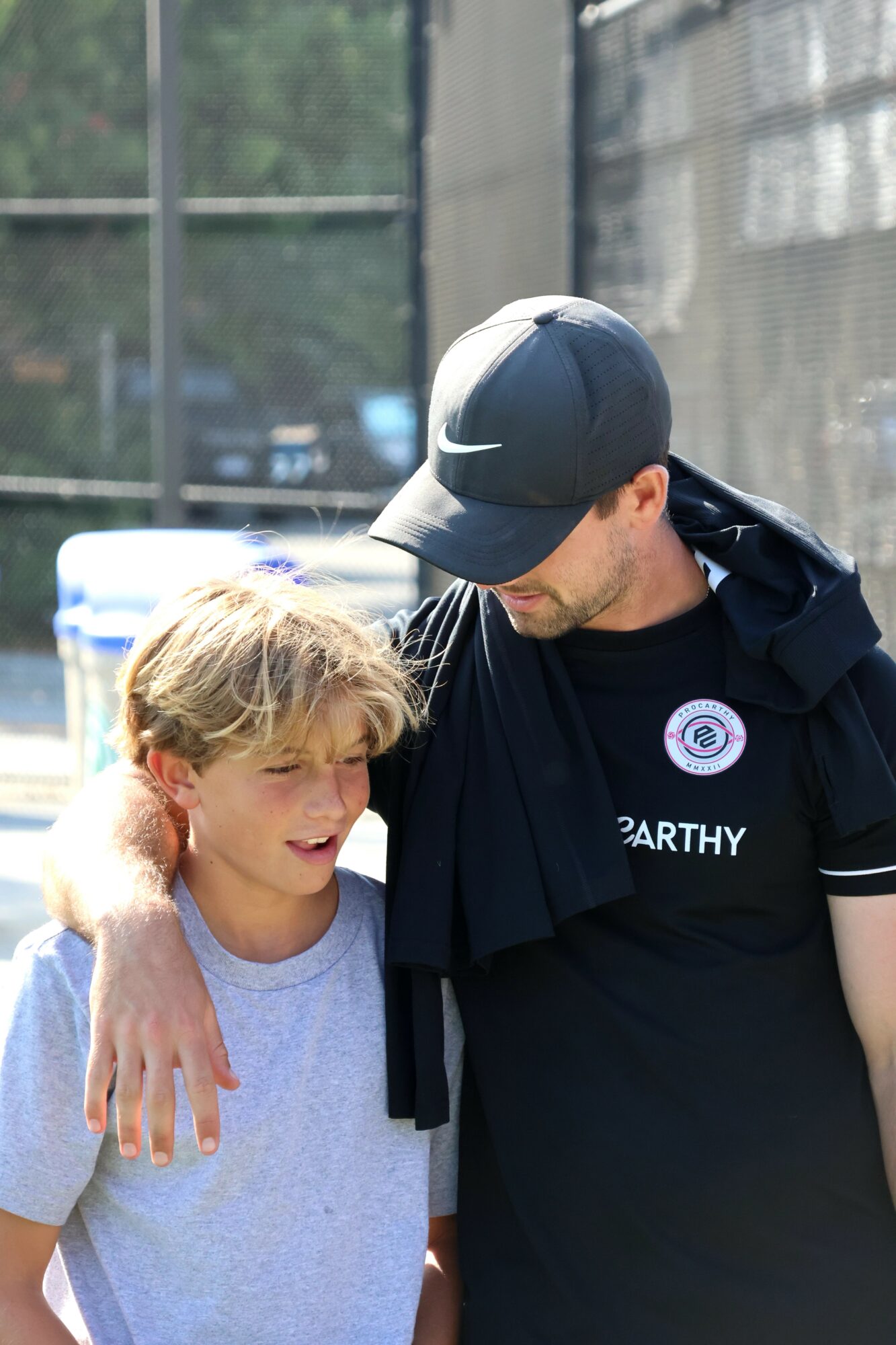 Two people, a young boy and an adult man, stand close together outdoors, with the man’s arm around the boy's shoulder.
