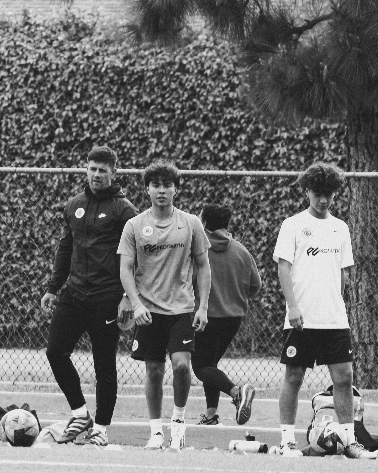 Four young men standing on a sports field with a fence and trees in the background.