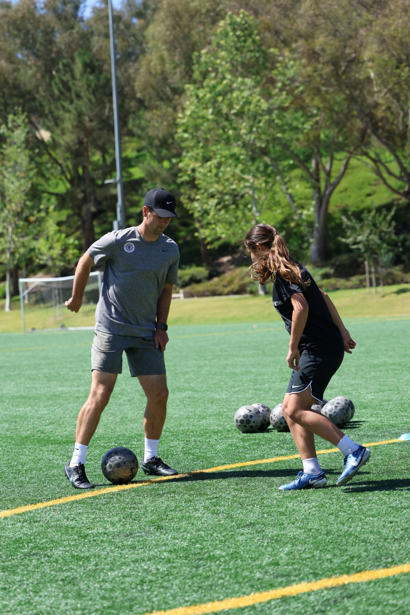 Two people on a soccer field with trees in the background, one person is about to kick a soccer ball, the other stands nearby.