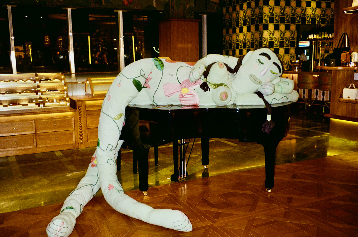 A large plush toy of a sleeping animal drapes over a black grand piano in a warmly lit room.