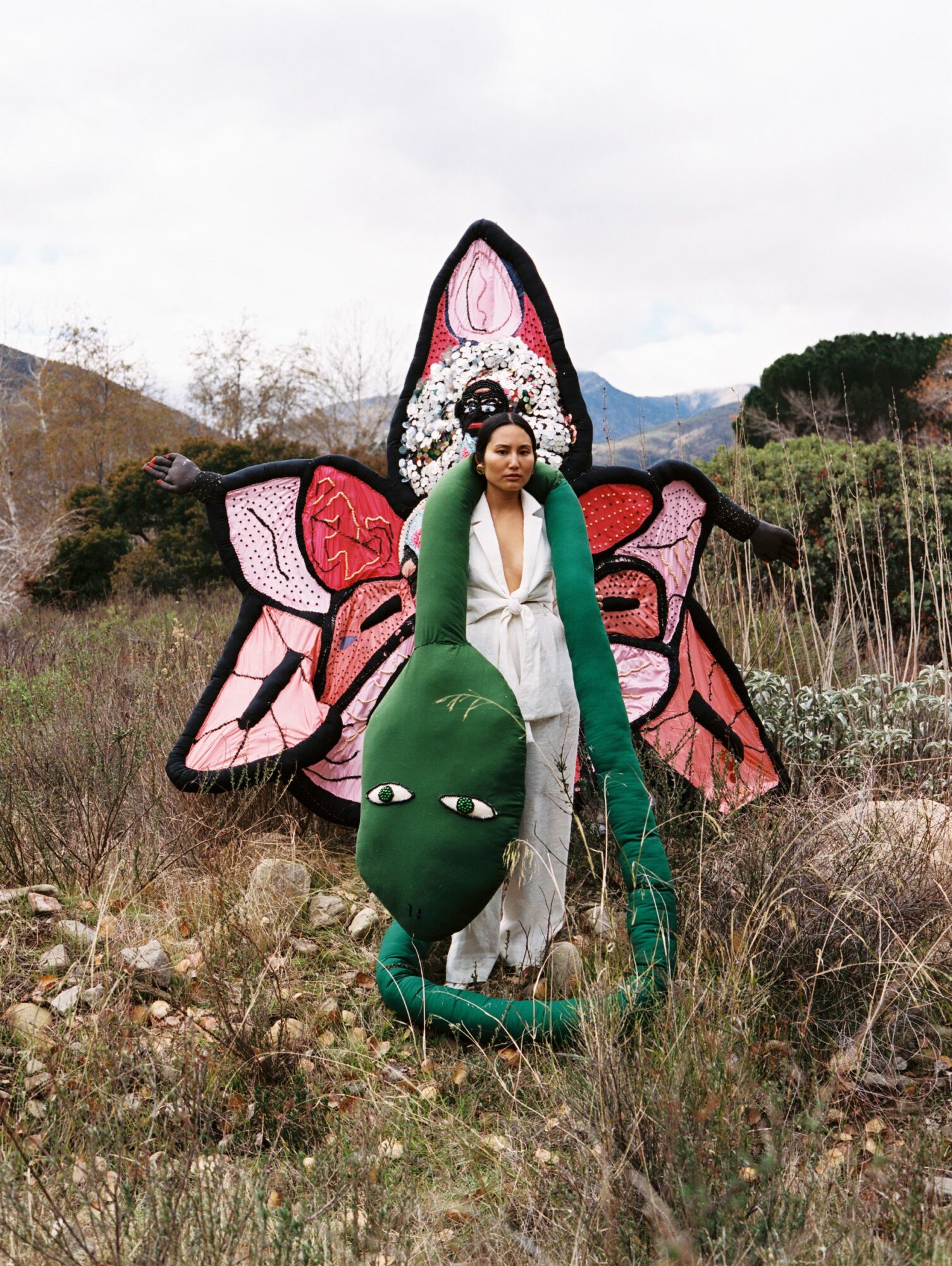 Person standing outdoors in front of a large butterfly-shaped kite with pink, black, and white colors, surrounded by grass and rocks.