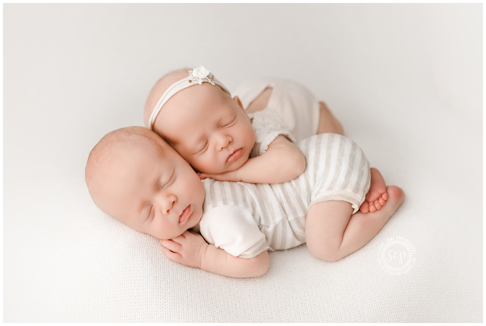 Two sleeping babies lying close together on a white surface, one with a headband, both with arms and legs relaxed.