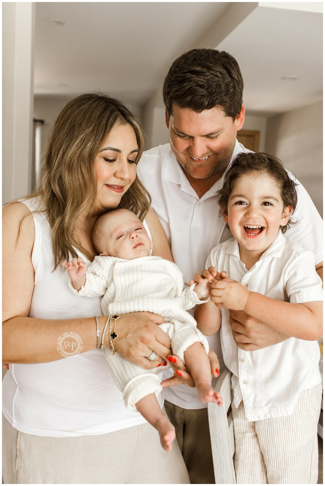 Family of four with a woman, man, and two children, smiling indoors, woman holding a baby, boy laughing.