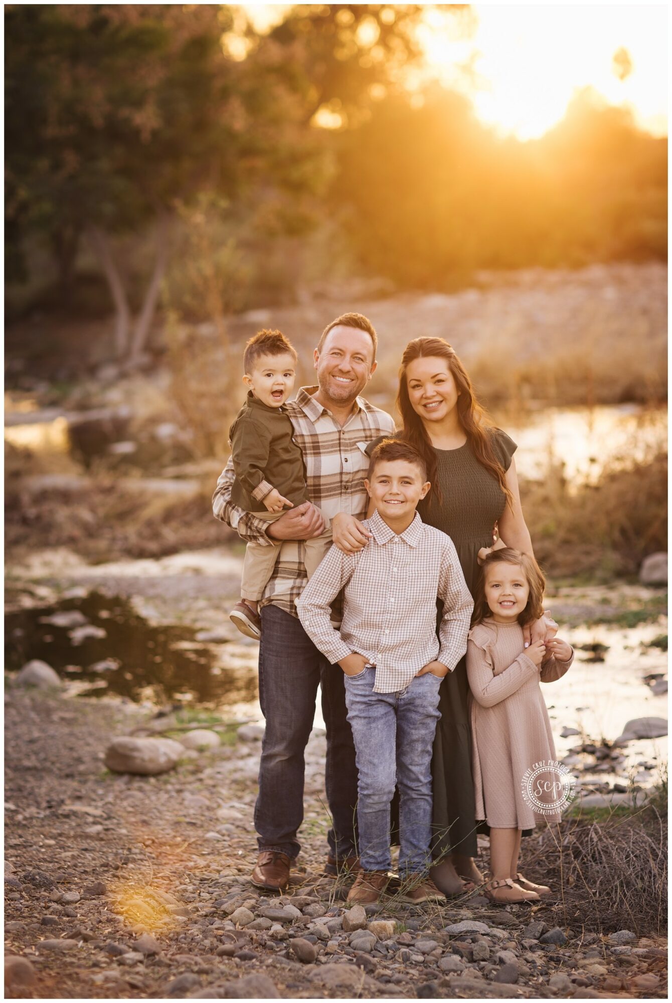 Family of five outdoors near water during sunset, smiling, standing close together, with trees in background.