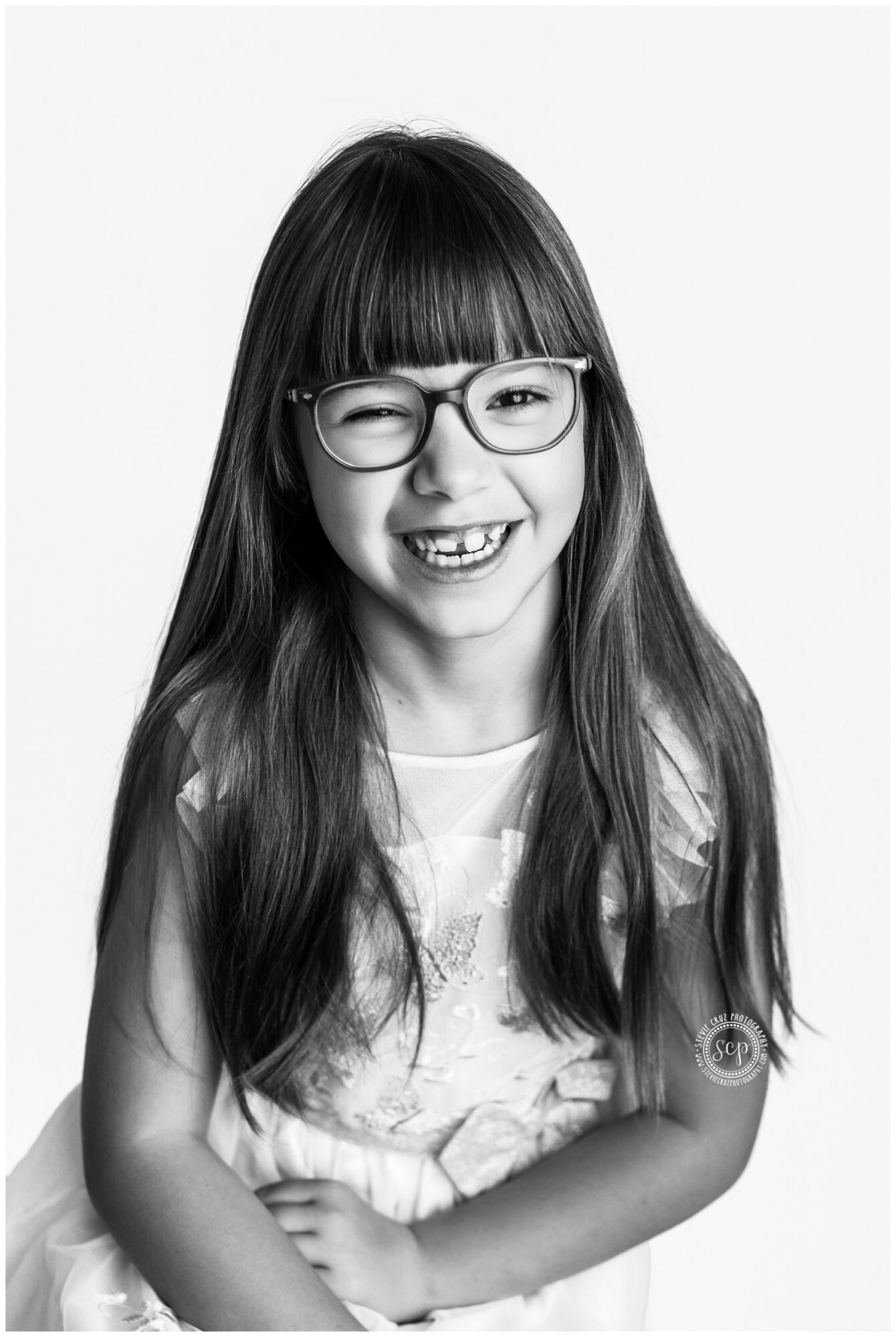 Young girl with long hair, glasses, smiling, wearing a light-colored shirt, standing against a plain background.