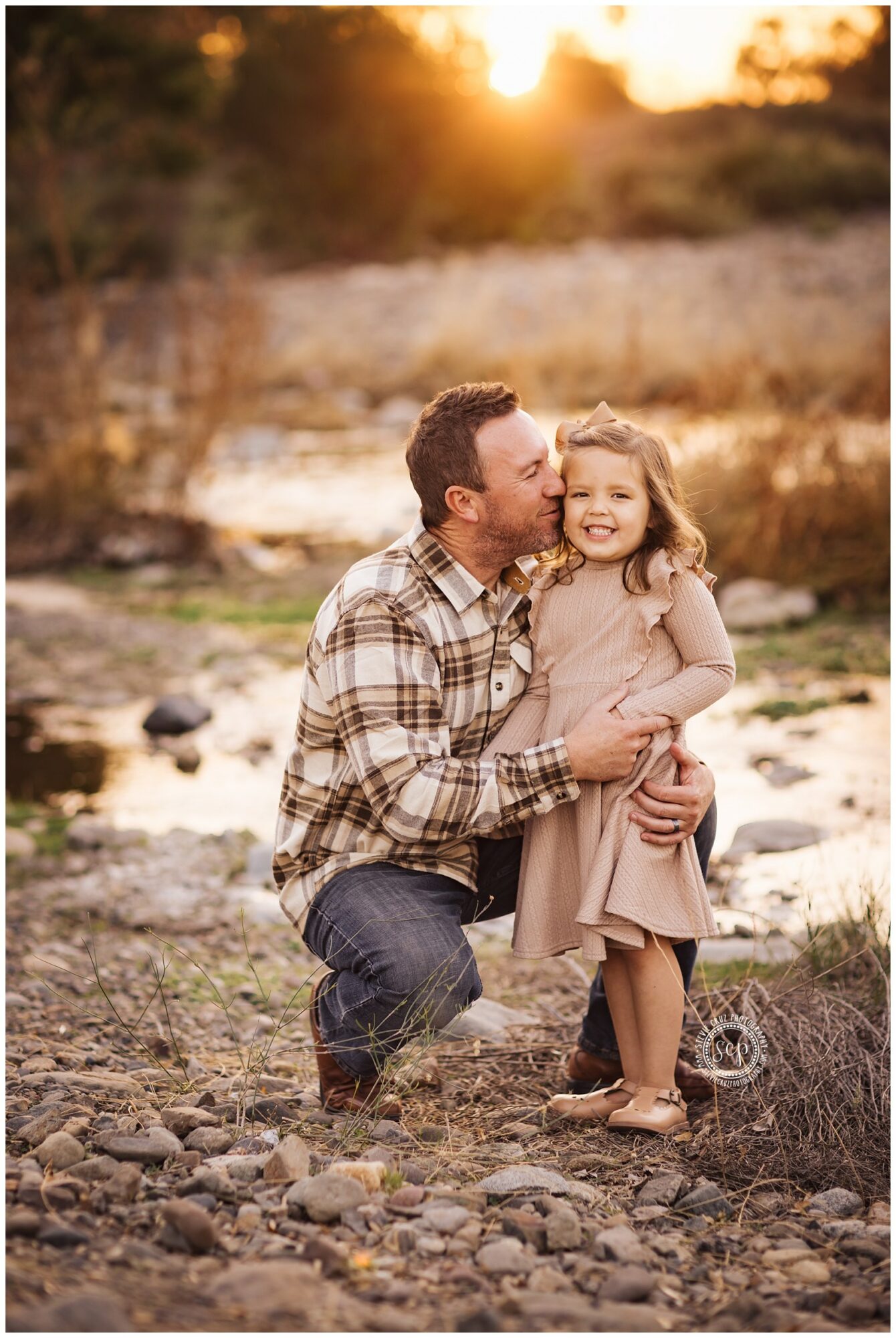 A man kneels beside a young girl outdoors during sunset, smiling and holding her hands, with a natural landscape background.