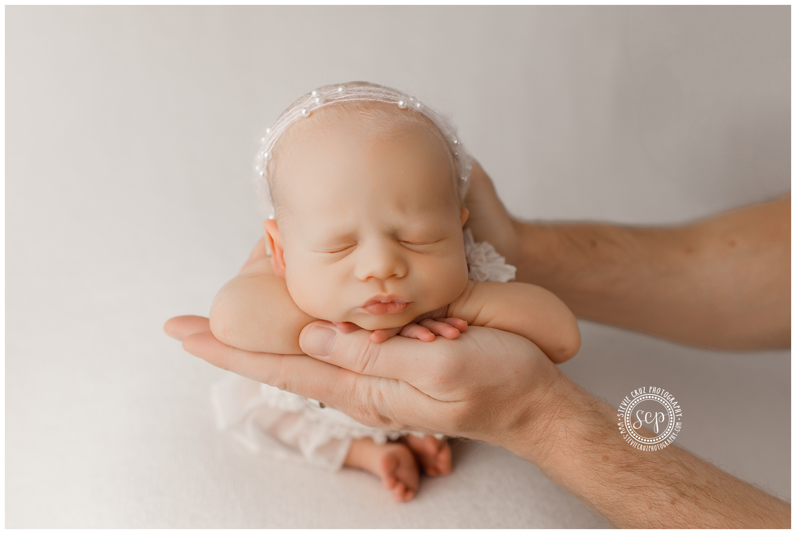Baby with closed eyes resting on hands, head supported, soft background, gentle lighting.