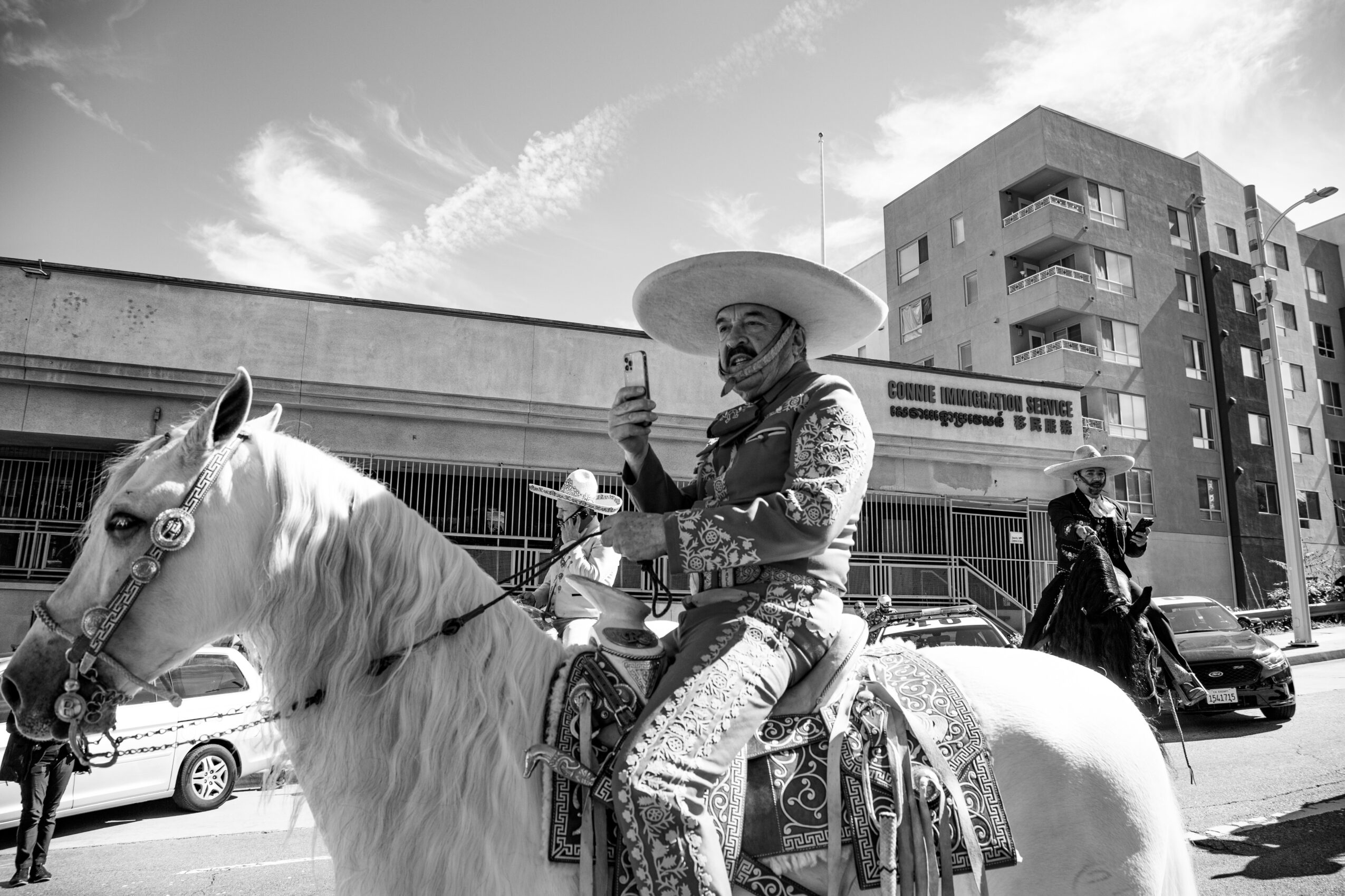 A person dressed as a mariachi with a large sombrero riding a white horse on a city street, with buildings and cars in background.