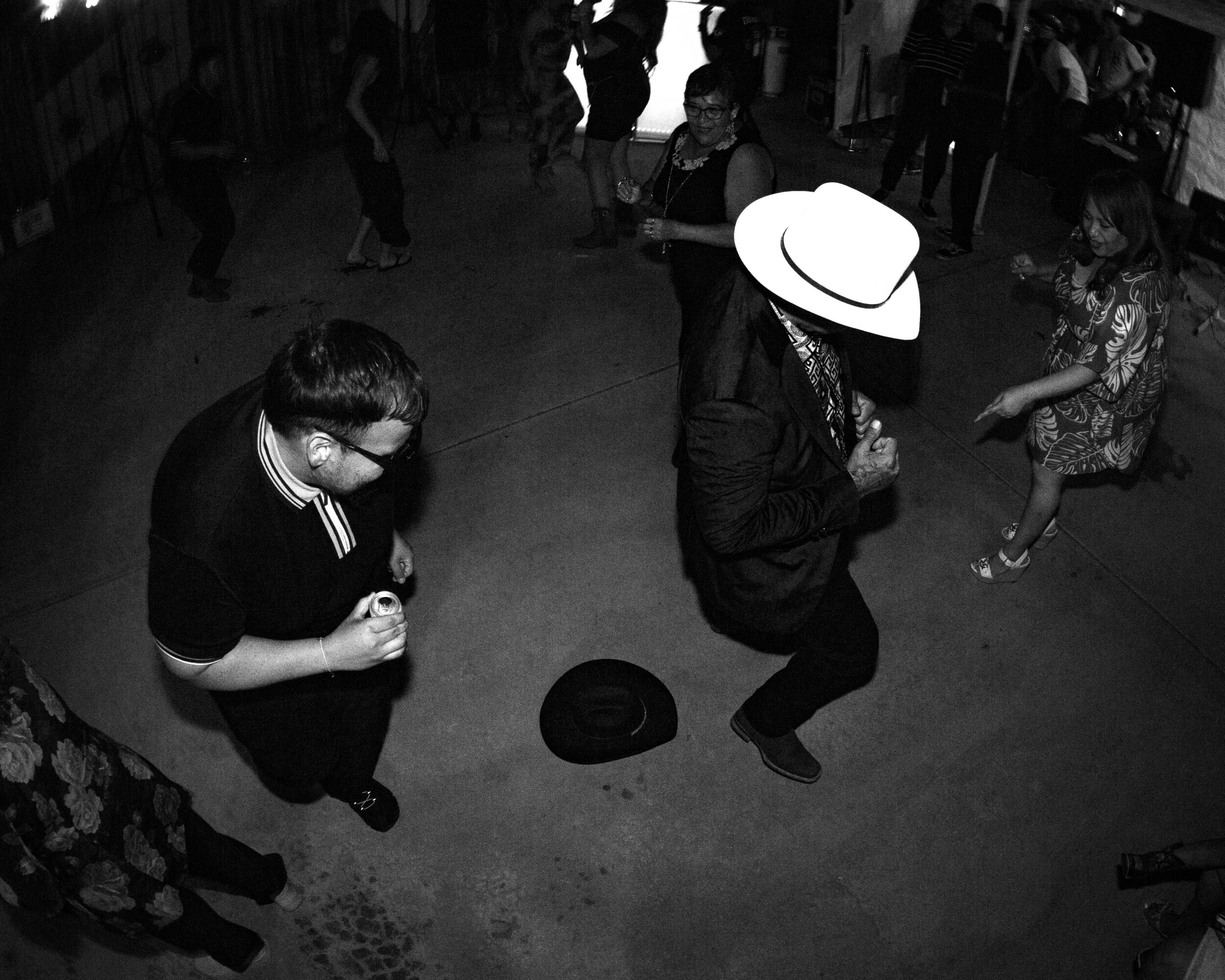 People dancing in a room, with one person wearing a large white hat, and a hat on the floor, in black and white.