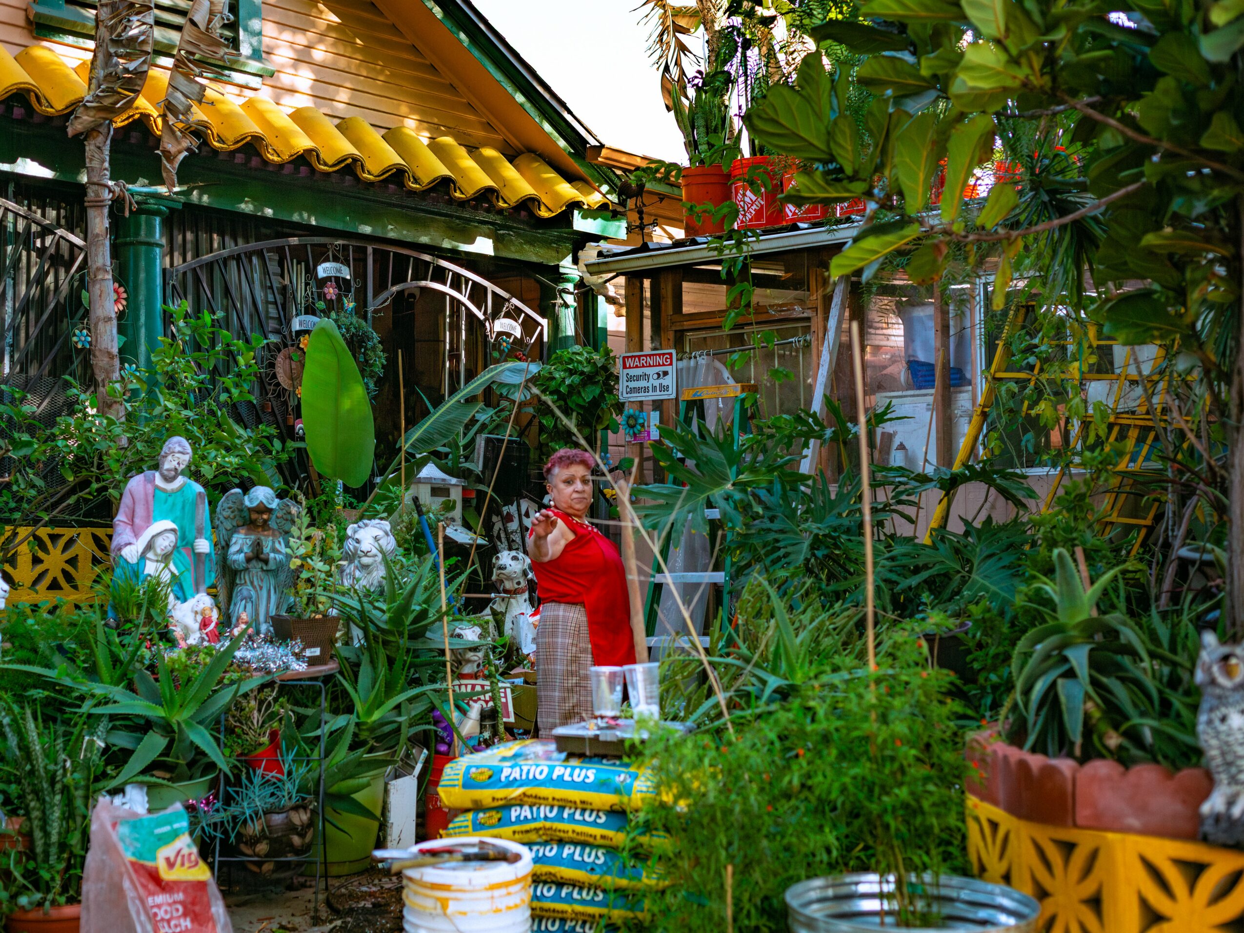 Child in red shirt standing among lush green plants and garden decorations in a vibrant outdoor setting.