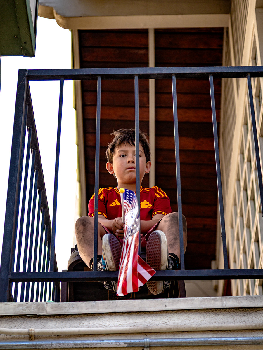 Child sitting on a balcony with a railing, holding an American flag, with a wooden ceiling above.