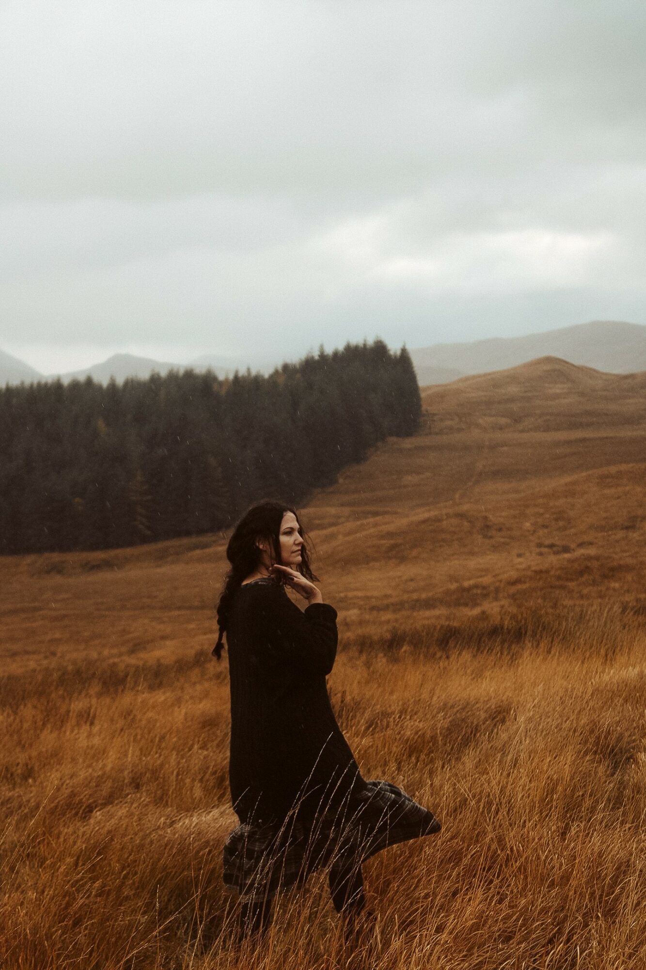 Woman standing in a grassy field with hills and cloudy sky in the background.