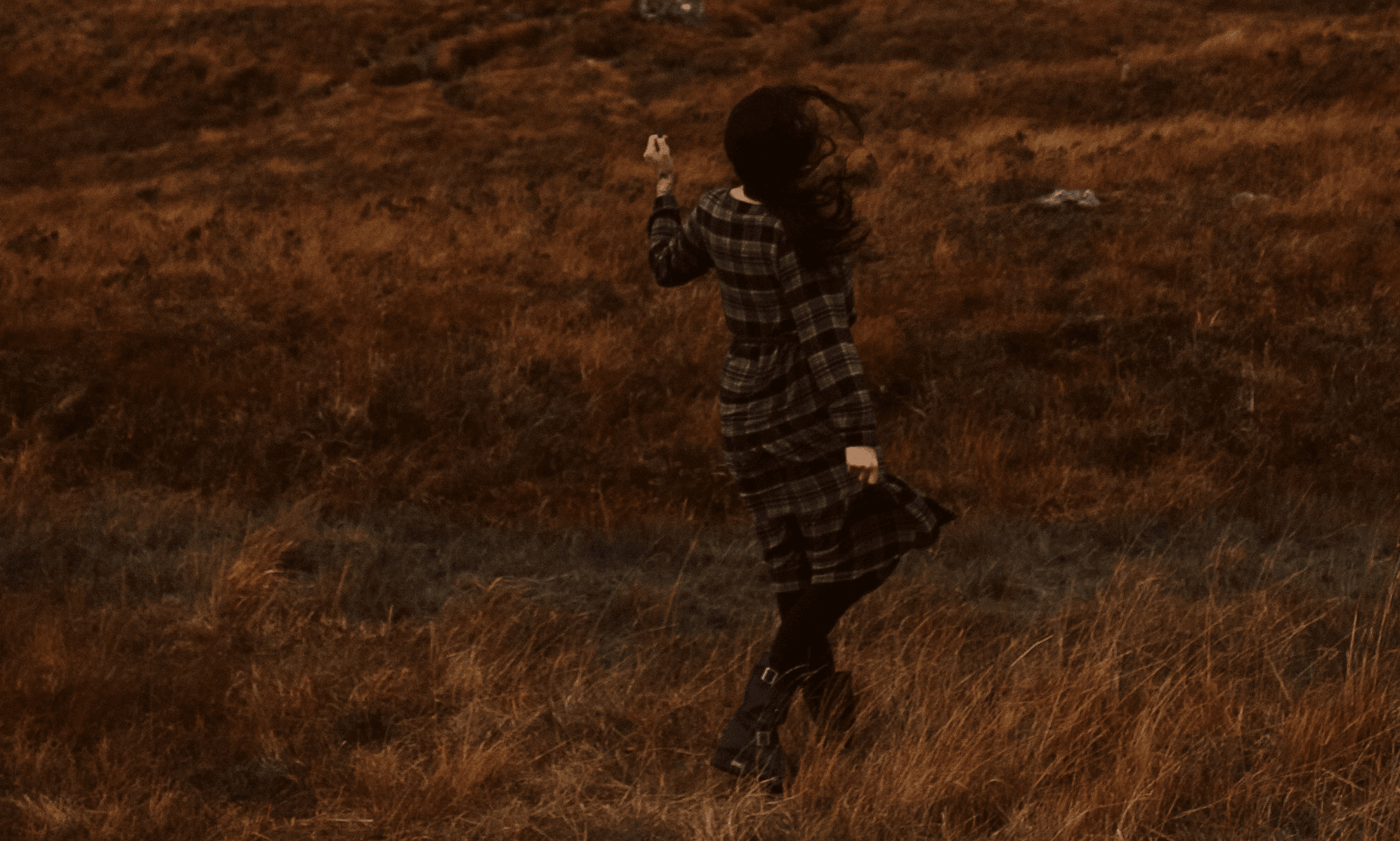 Person standing in a grassy field, holding a phone, with a landscape background, facing away from the camera.