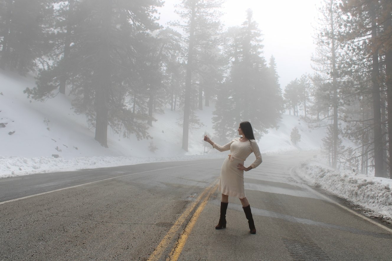 Woman in a white dress and boots standing in the middle of a foggy, snow-covered road surrounded by trees.