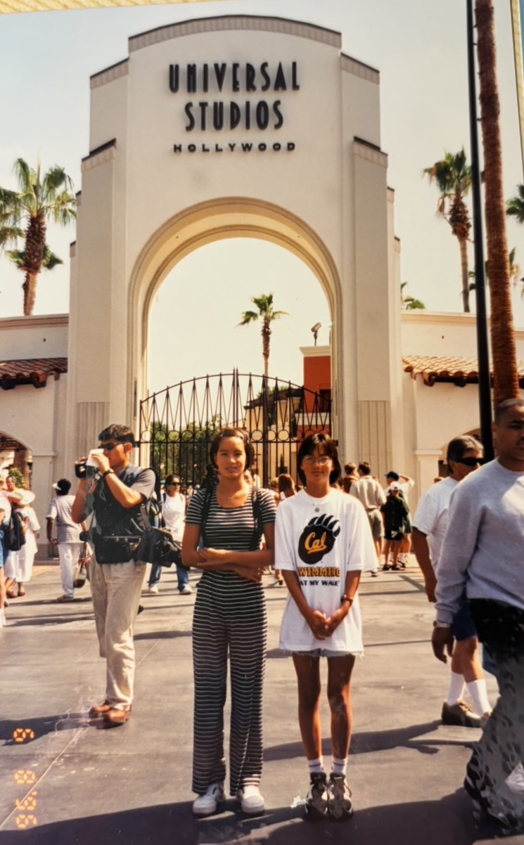 People walking outside the entrance of Universal Studios Hollywood, with a large archway and palm trees in the background.