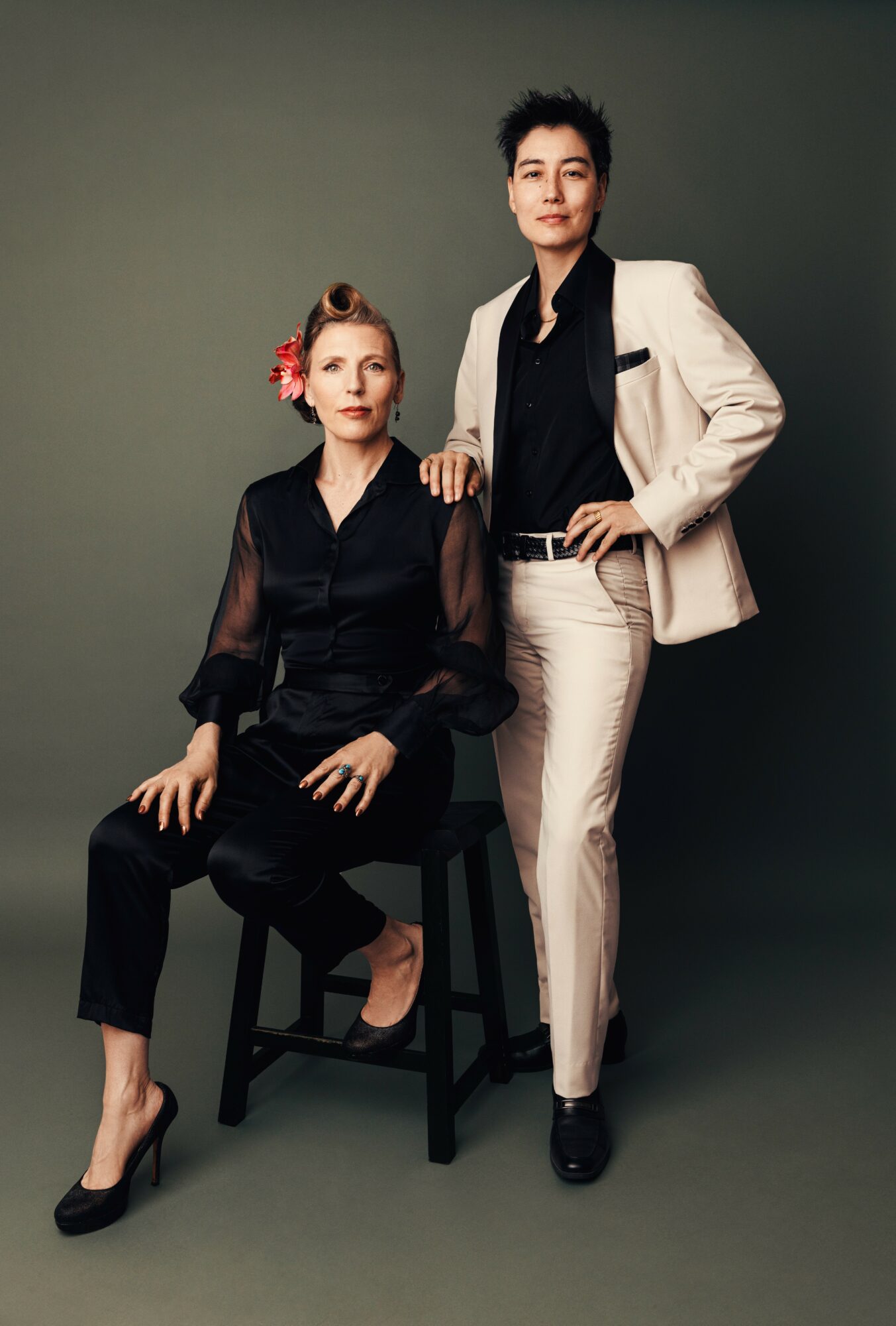 Two women pose against a plain background, one seated on a stool and the other standing beside her, both dressed stylishly.