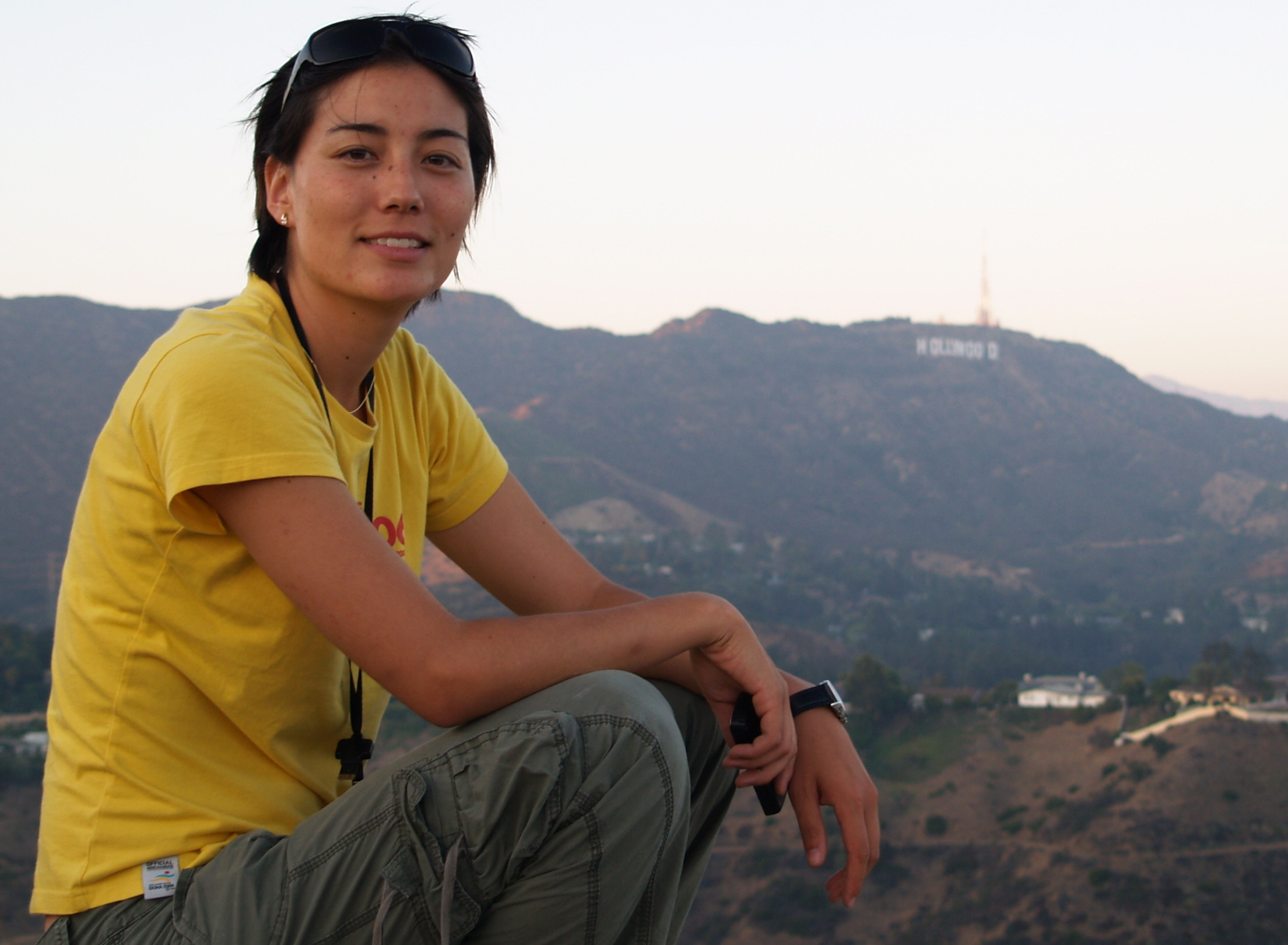 Young woman sitting outdoors with mountains in the background, wearing a yellow shirt and gray pants.