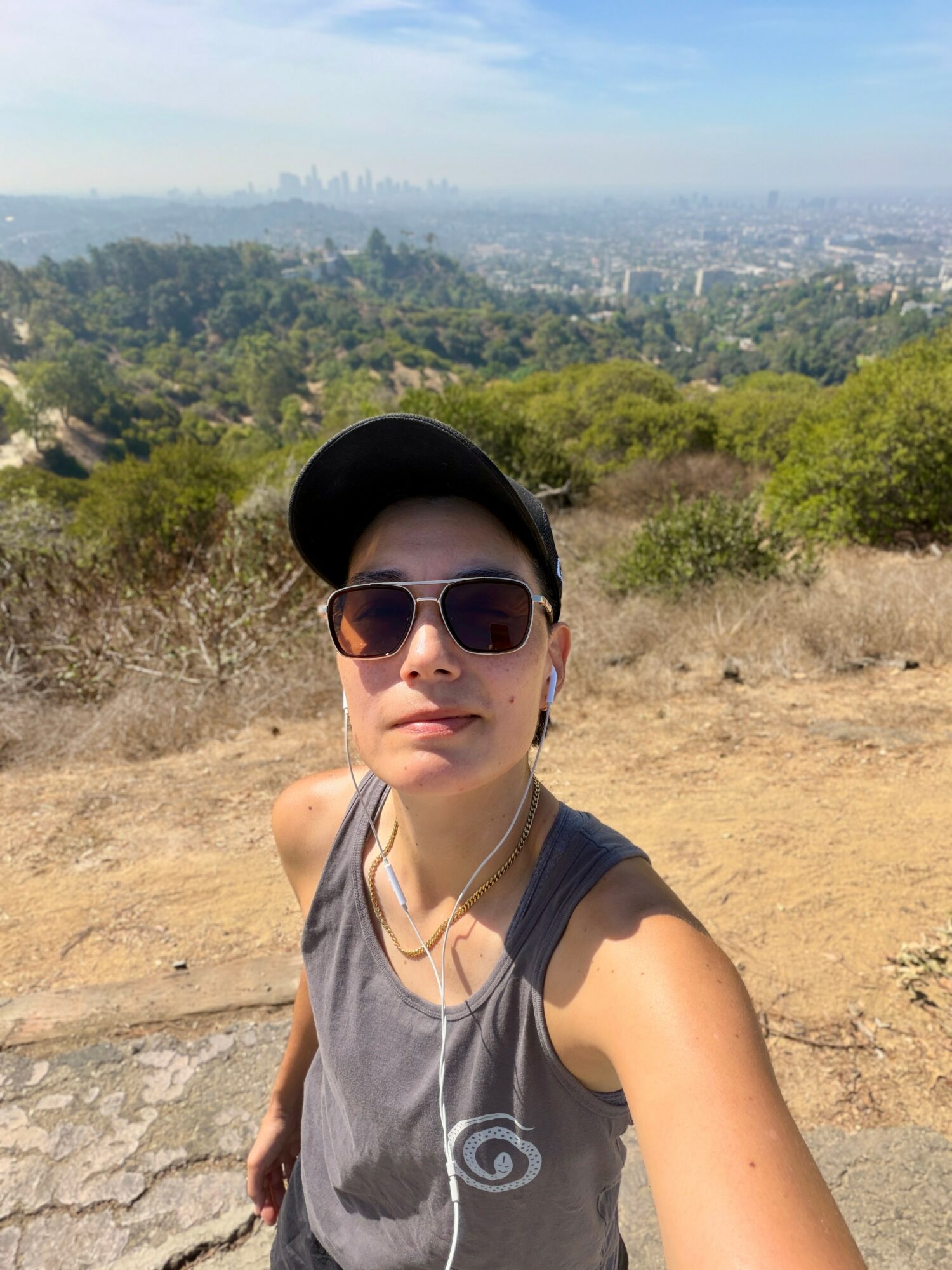 Person wearing sunglasses and a cap taking a selfie outdoors with a cityscape in the background.