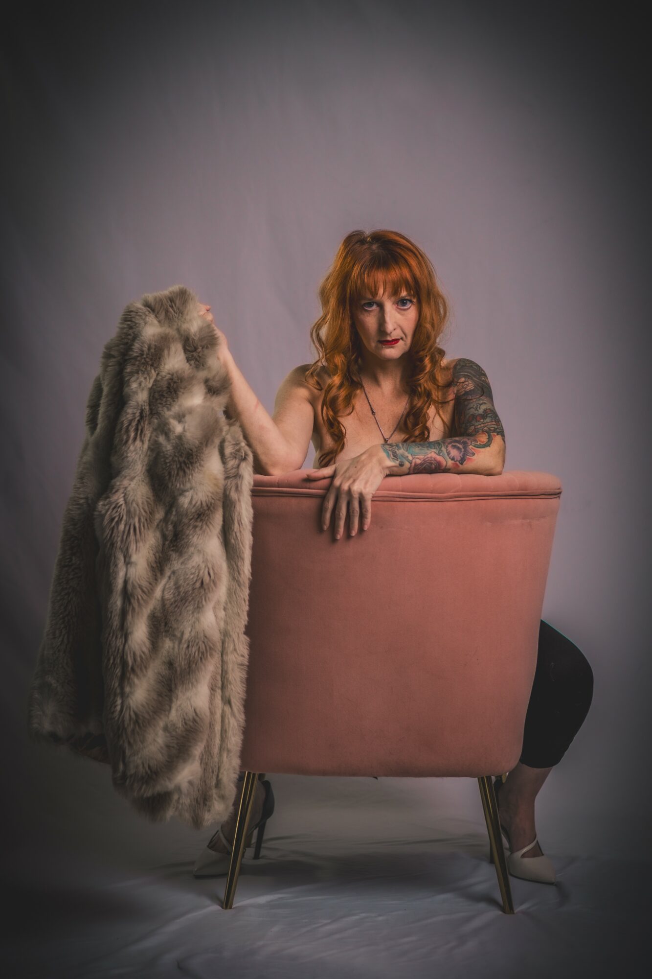 Woman with red hair and tattoos leaning on a pink chair, with a fur coat draped over the back, against a plain background.