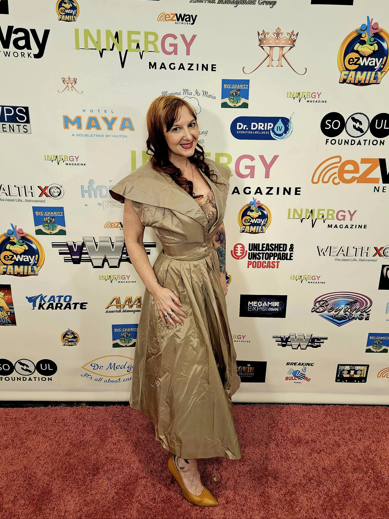 Woman in a beige dress standing on red carpet in front of a backdrop with various logos and text.