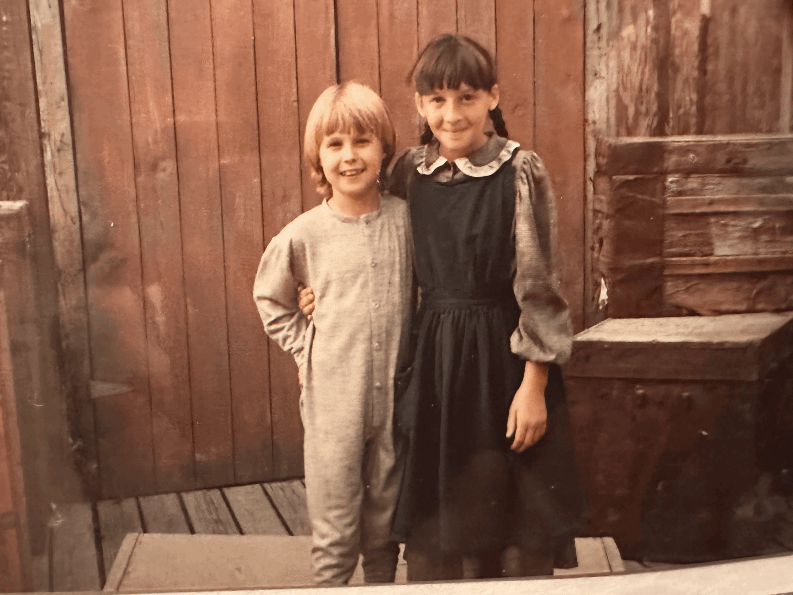 Two children, a boy and a girl, stand side by side outdoors in front of a wooden fence, smiling.