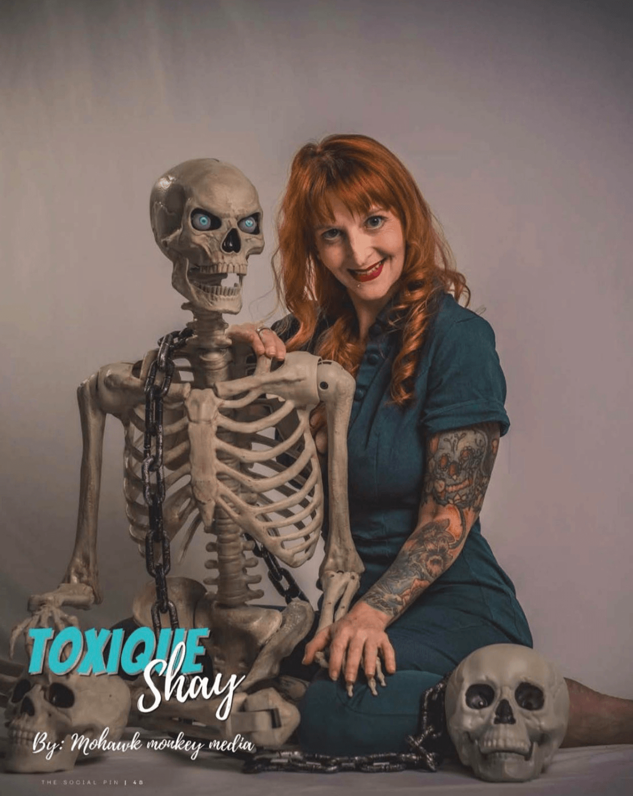 Woman with red hair and tattoos sitting next to a skeleton with a chain, smiling, in a studio setting.