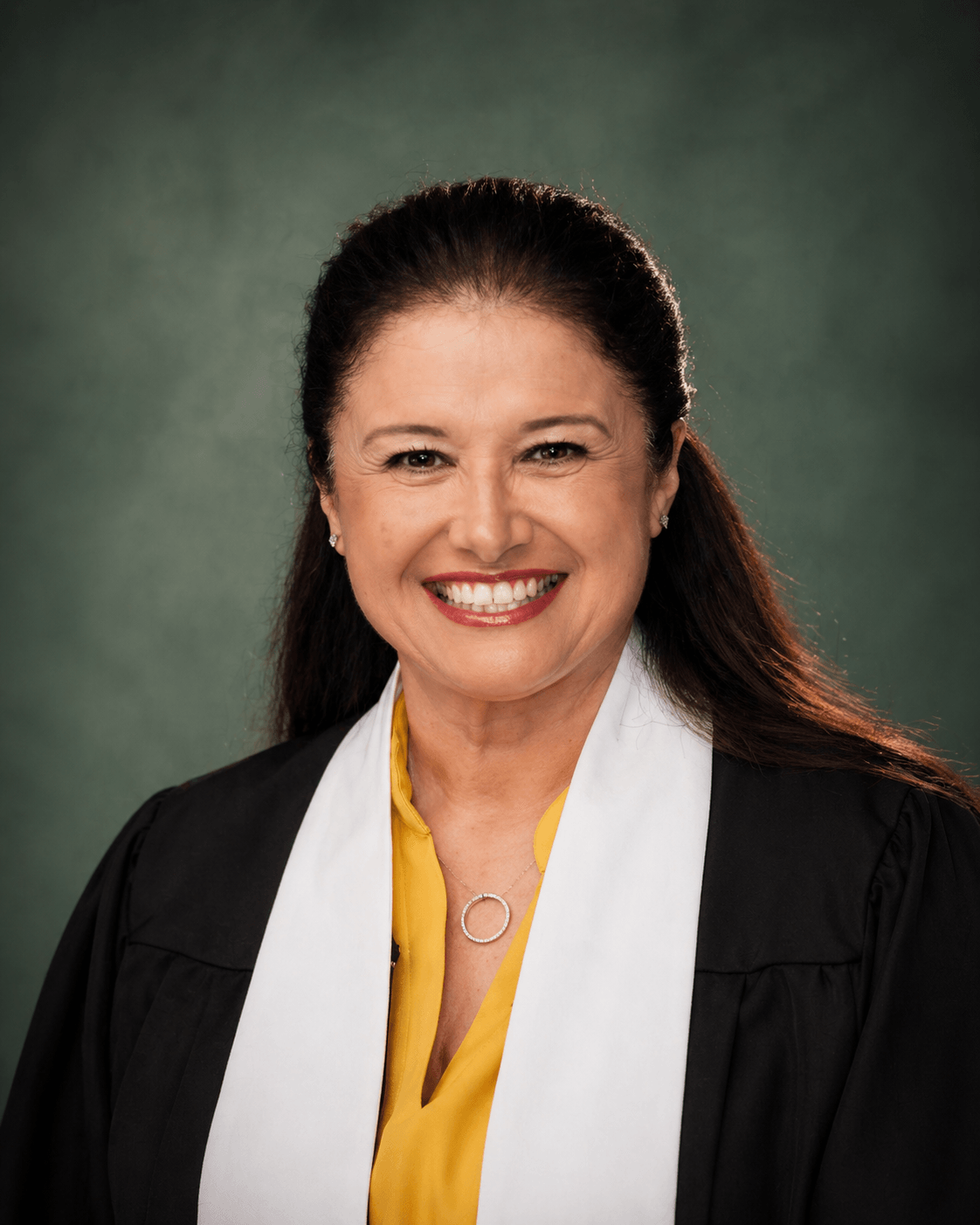 Smiling woman with long dark hair, wearing a black robe, white collar, and yellow blouse, against a green background.