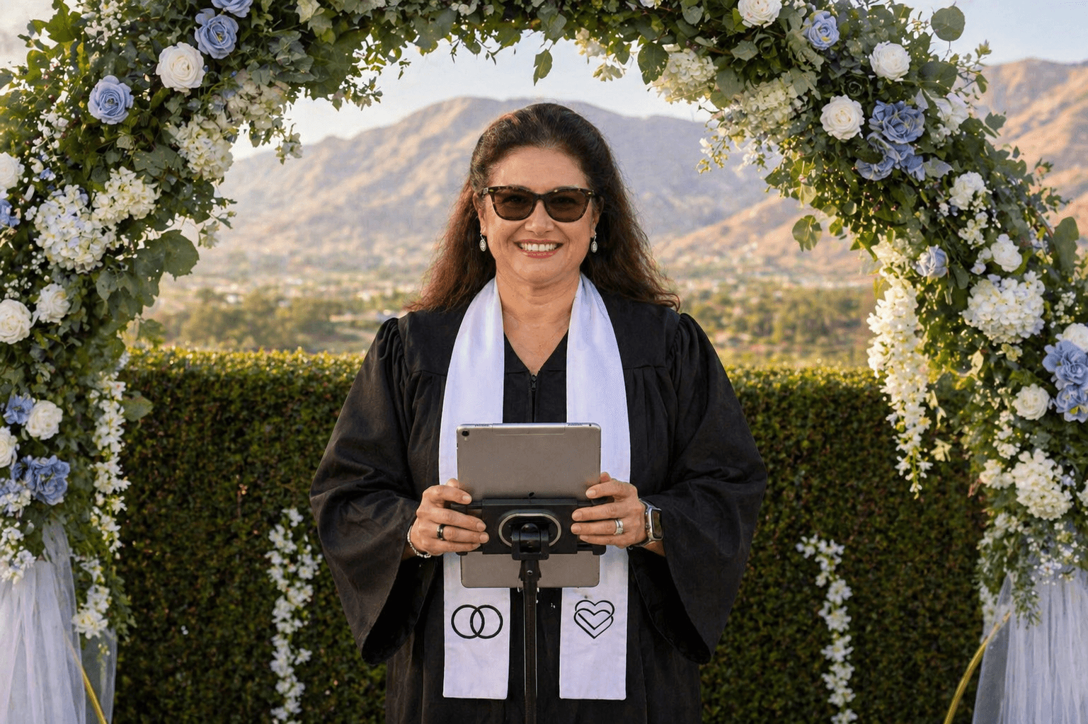 Woman in graduation gown holding a tablet, standing under a floral arch outdoors, smiling, mountains in background.