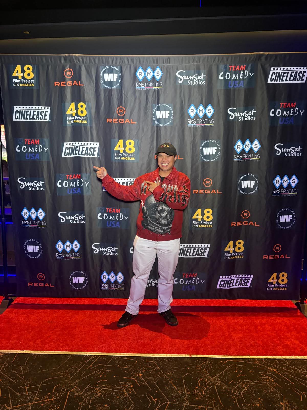 Person standing on red carpet in front of a backdrop with logos, wearing a red sweater, white pants, and a cap, smiling and pointing.