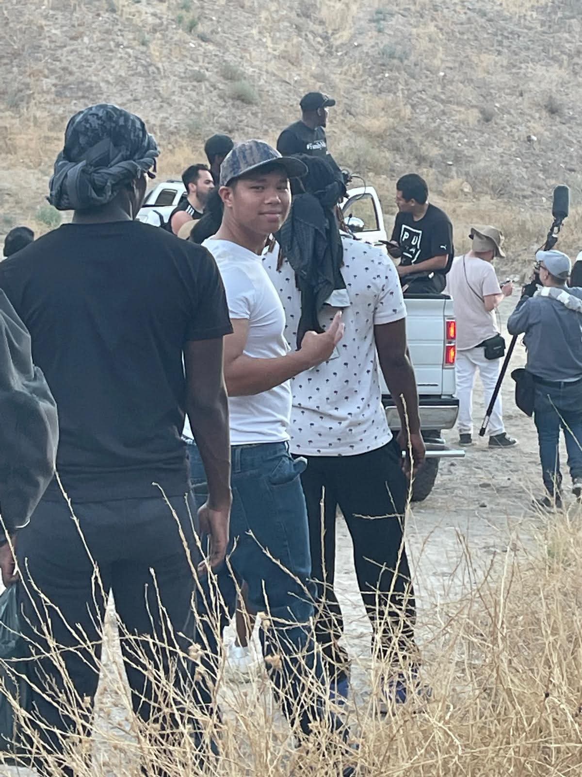Group of people outdoors in a dry, grassy area with vehicles and a rocky background, some wearing hats and backpacks.