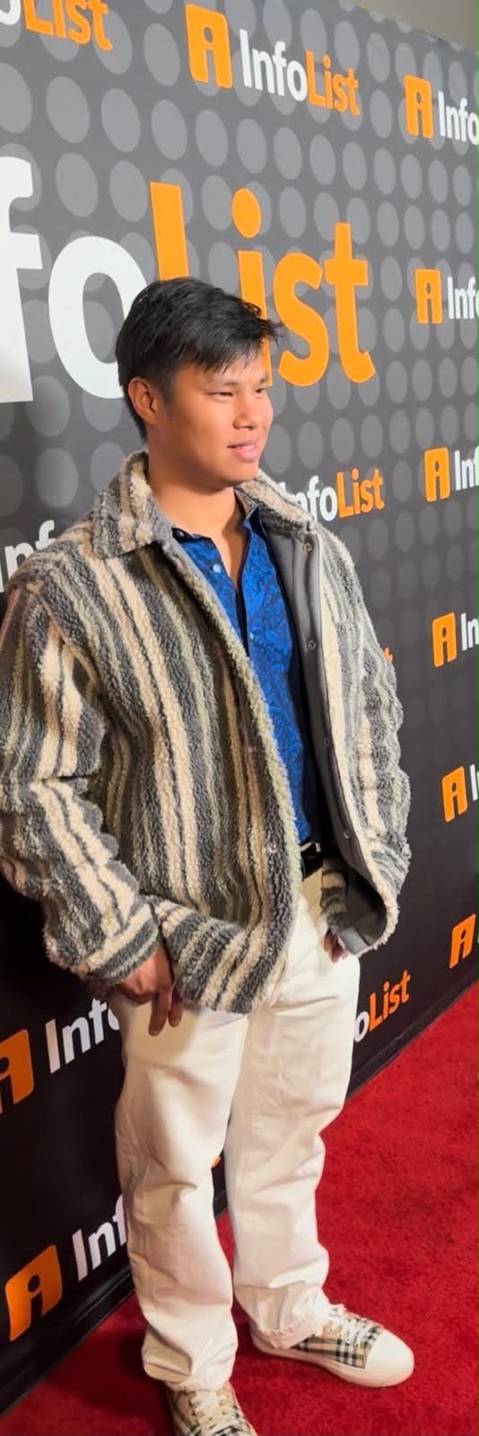 Young man with dark hair wearing a striped jacket, blue shirt, and beige pants, standing on a red carpet in front of a black backdrop with orange and white text.