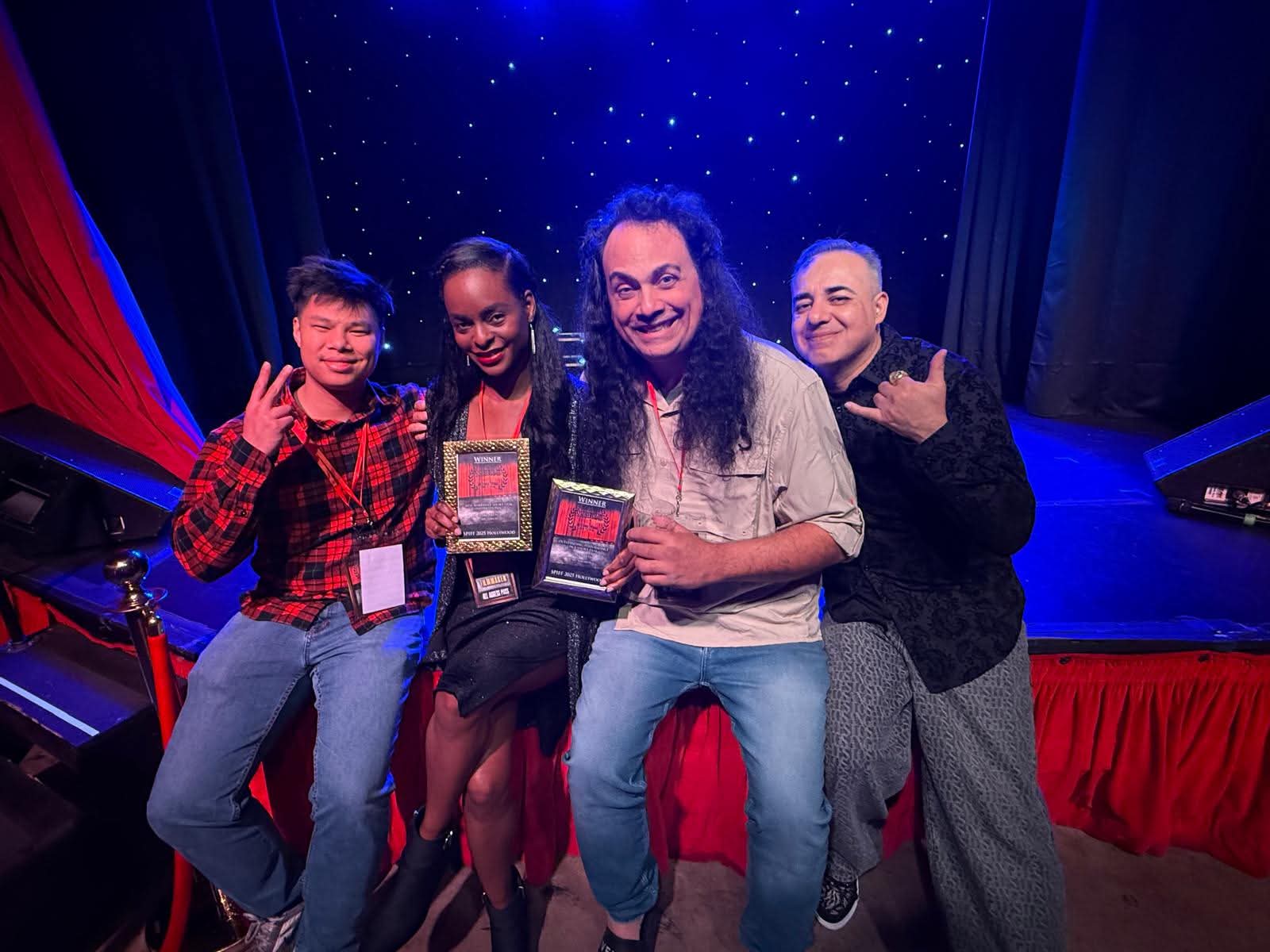 Four people smiling and posing on stage with a starry background, some making hand gestures, in a group photo.