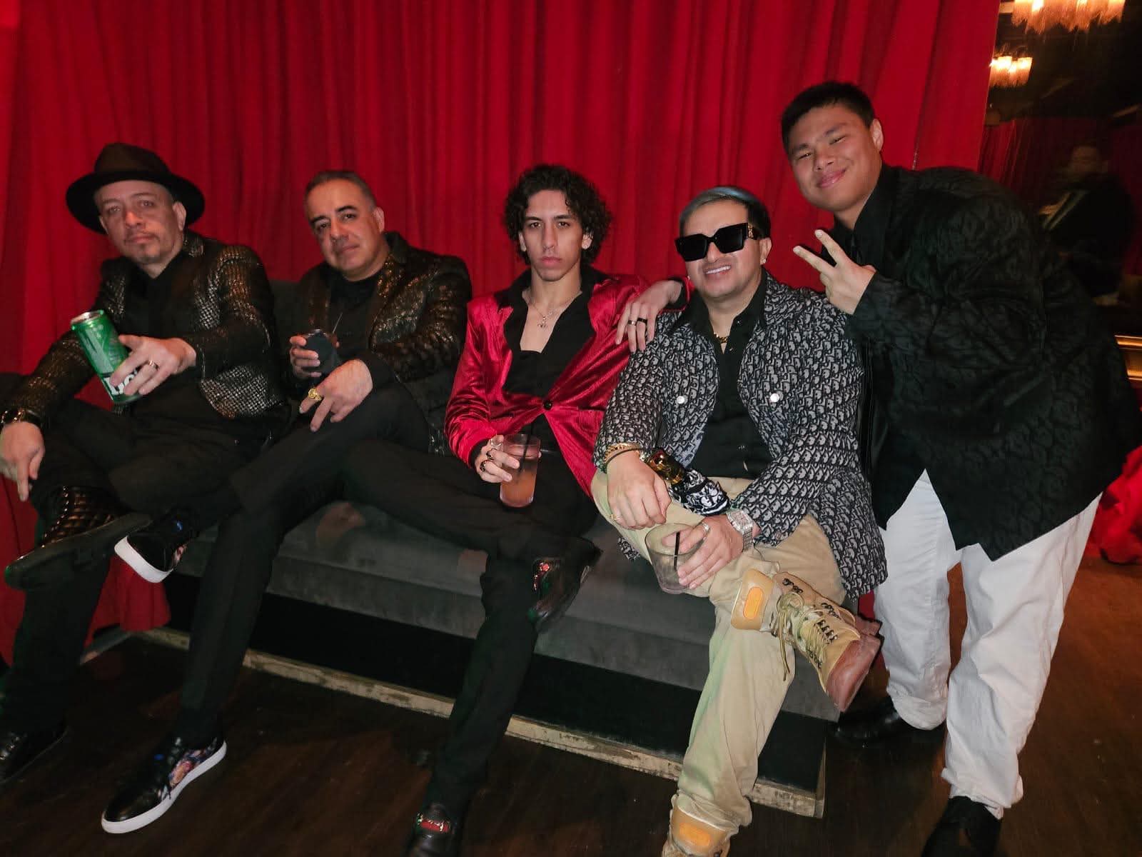 Six people sitting on a bench in front of a red curtain, posing for a photo, some wearing sunglasses, one making a peace sign.