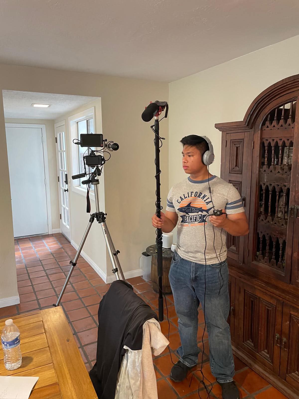 Young boy holding a boom microphone in a room with camera equipment, wooden cabinet, and a table with a water bottle.