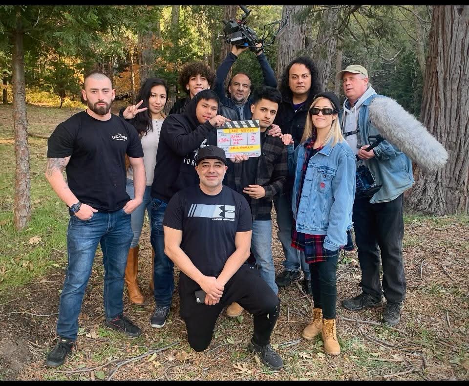 Group of ten people outdoors in a wooded area, some holding a clapperboard, posing for a photo.