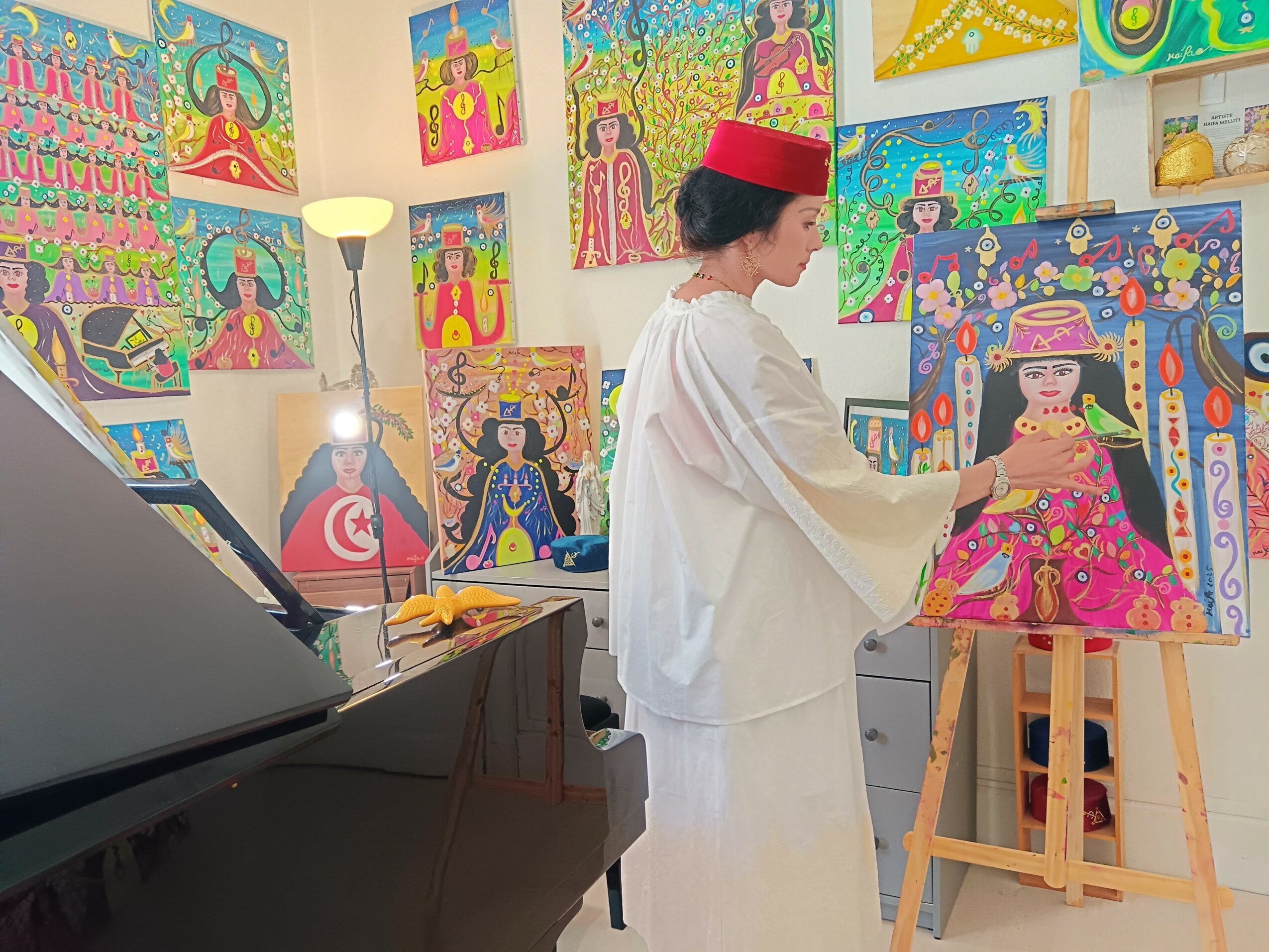 Person in white clothing and red hat painting a colorful artwork on an easel, surrounded by vibrant paintings on the wall.