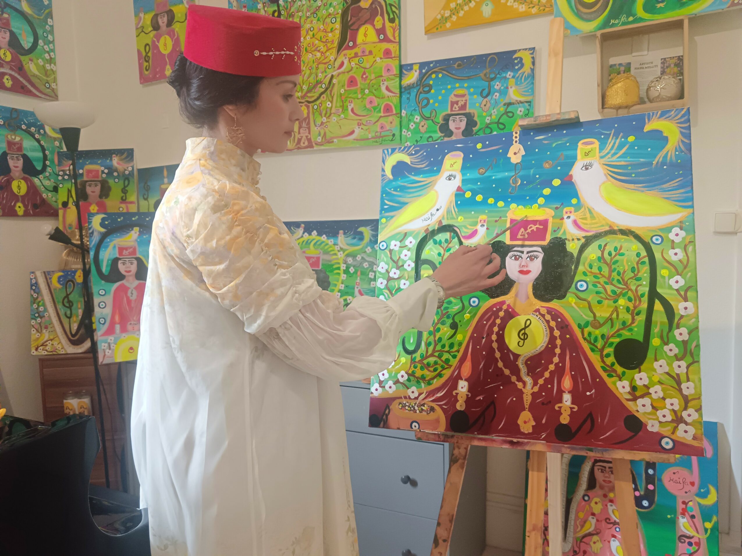 Woman in traditional attire painting colorful artwork on canvas, surrounded by other vibrant paintings, in an art studio.