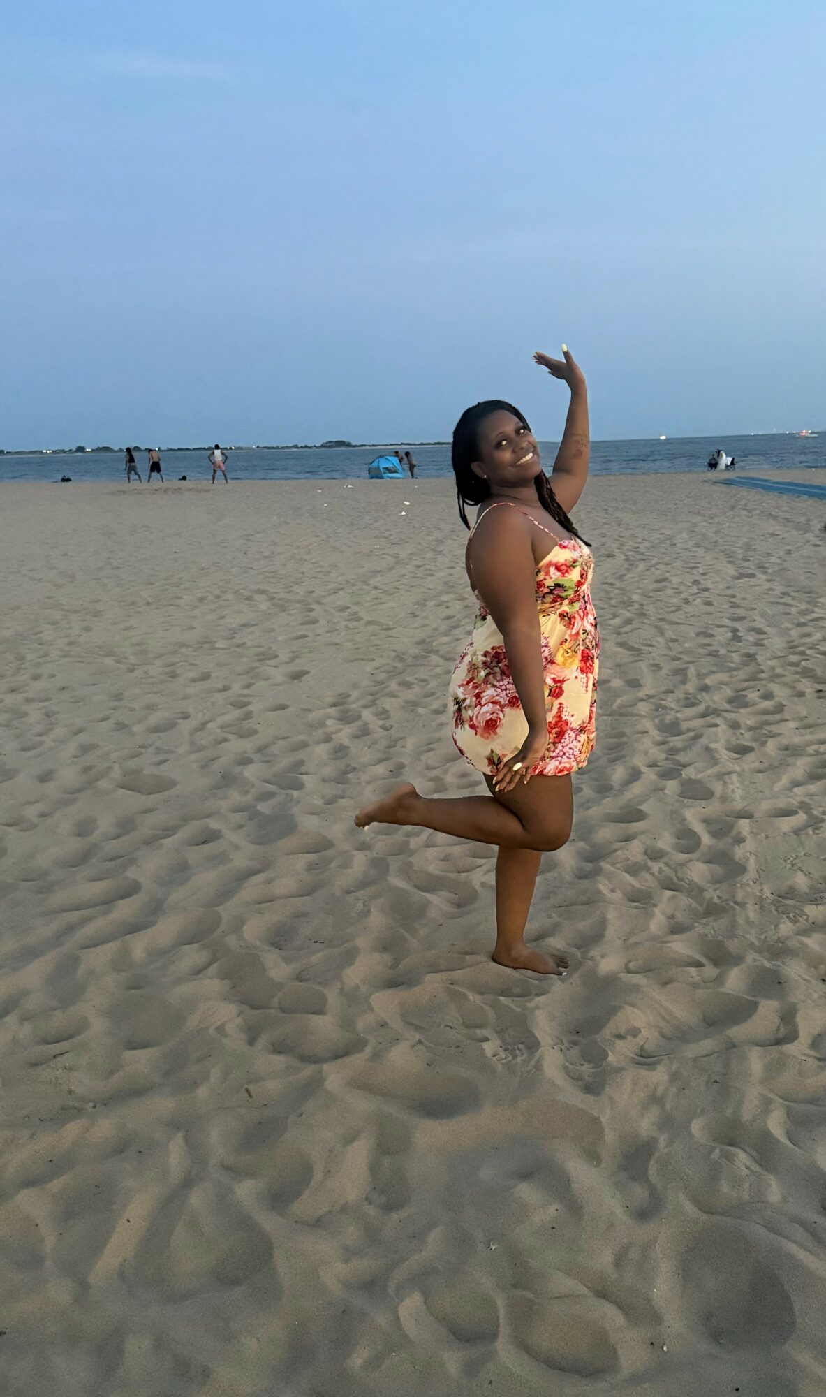 Woman in floral dress balancing on one leg on sandy beach, with ocean and sky in background.