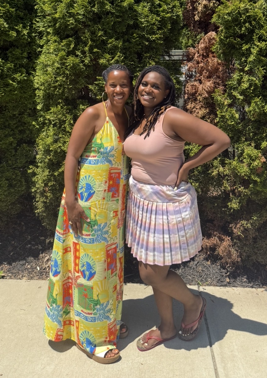 Two women standing outdoors in front of green and brown foliage, smiling, one in a colorful dress, the other in a pink top and striped skirt.