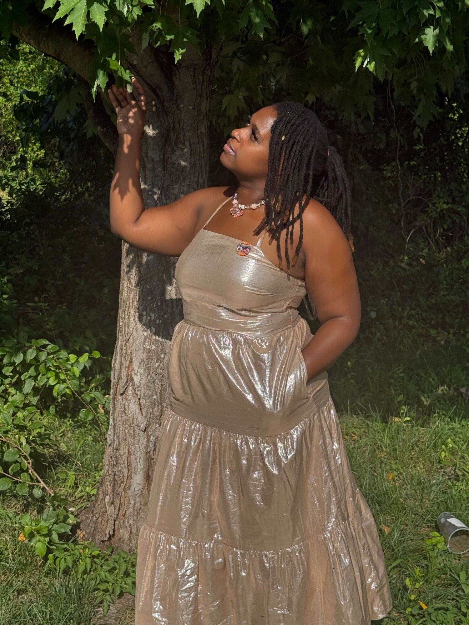 Woman with braided hair leaning against a tree in a beige dress outdoors, surrounded by greenery.