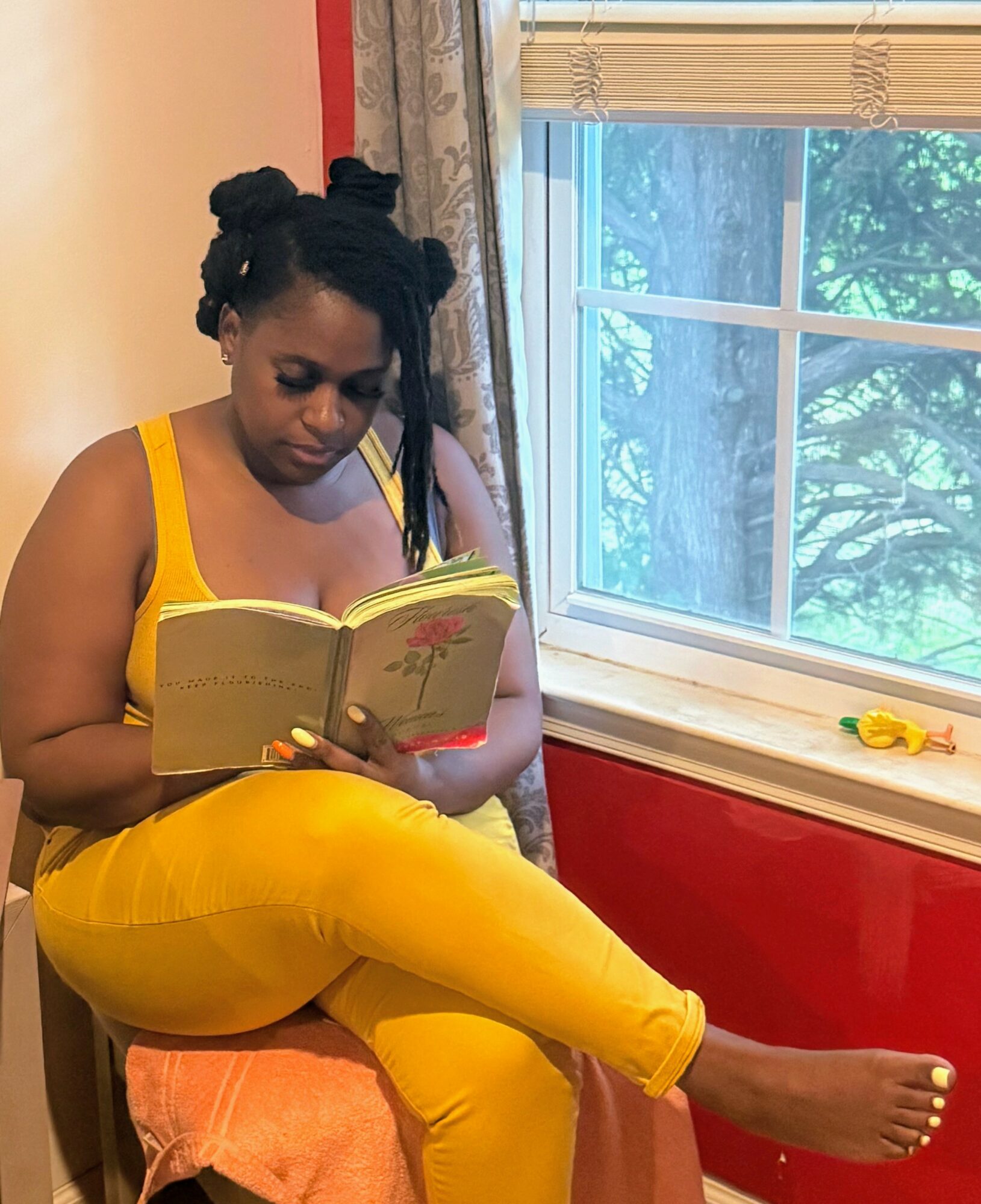 Woman in yellow outfit reading a book while sitting on a chair near a window with trees outside.
